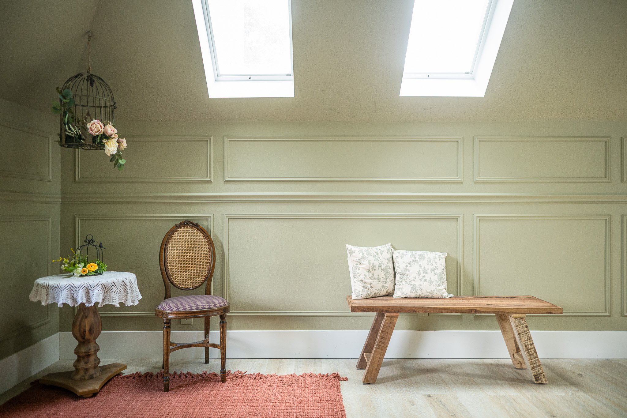 A cozy attic room with light green paneled walls, two skylights, a small round table with a lace tablecloth and yellow flowers, a wooden chair with a woven backrest and purple-striped cushion, a rustic wooden bench with two floral pillows, a pink rug