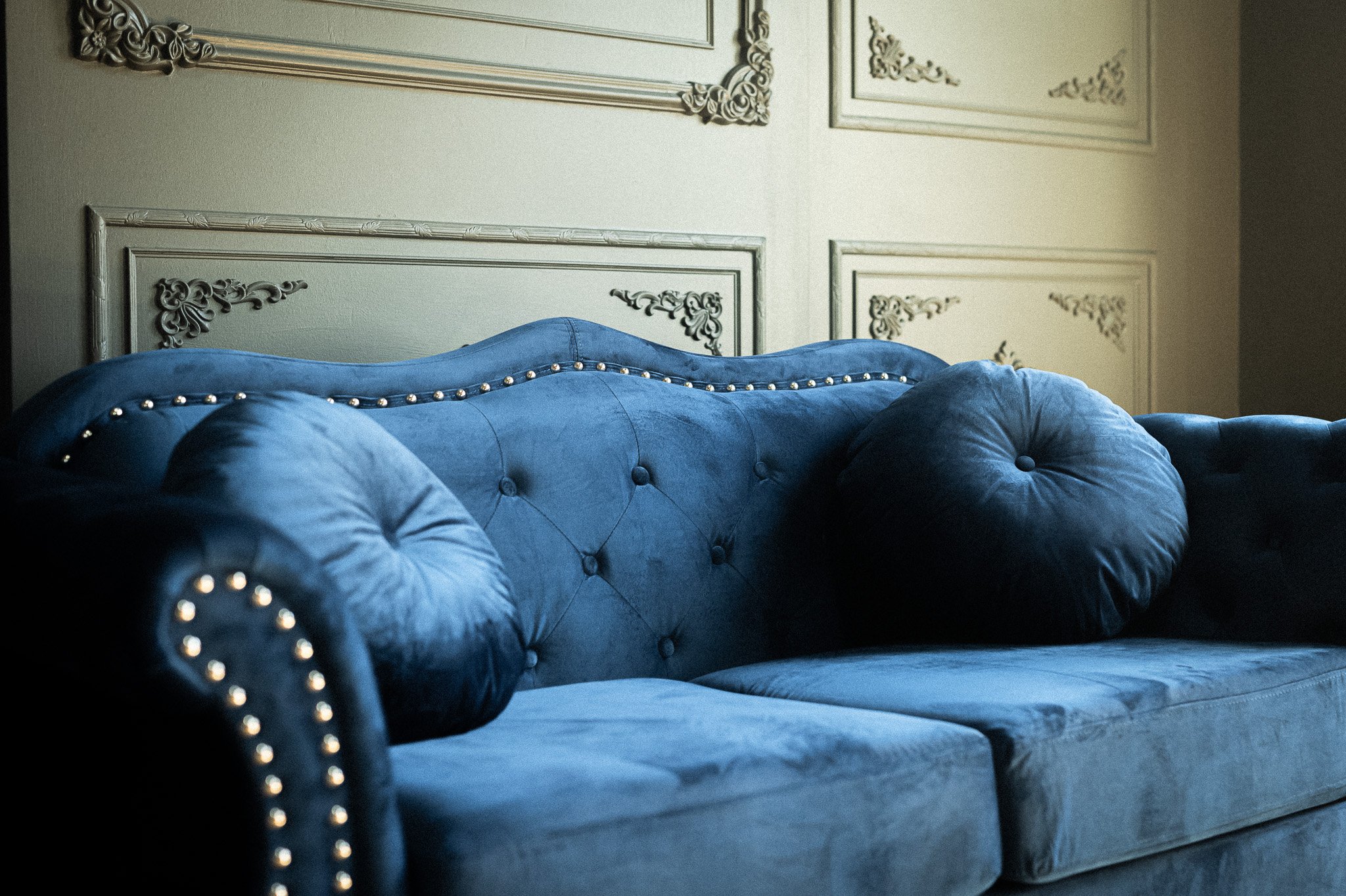 A blue velvet tufted sofa with rolled arms and gold nailhead trim in a room with ornate grey wall paneling.
