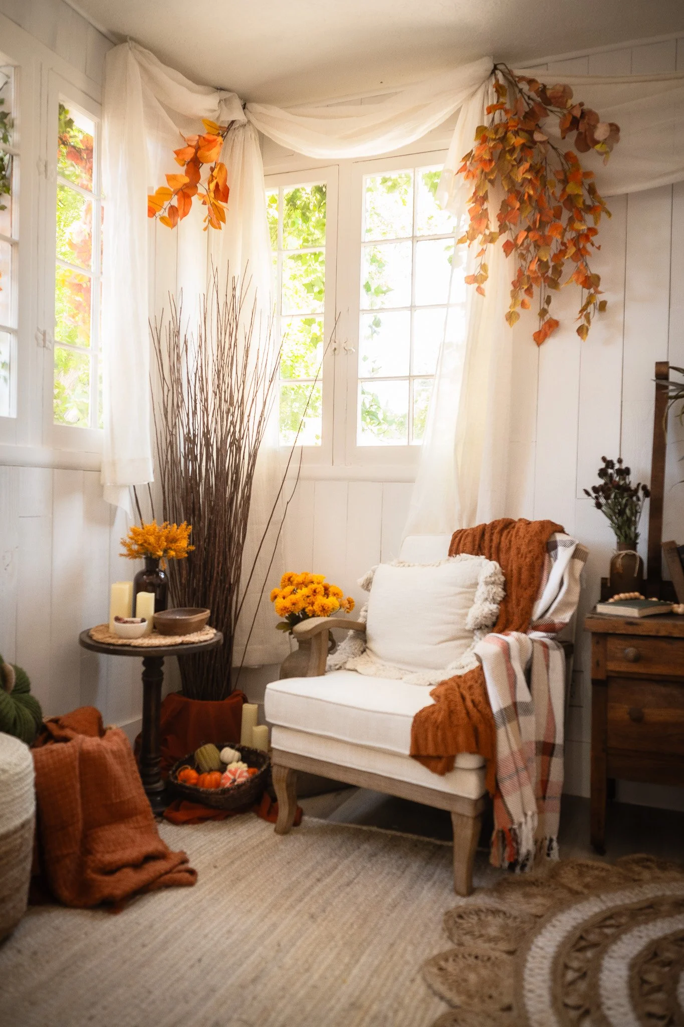 A cozy corner of a room with a white armchair, autumnal decor, and sunlight streaming through windows.