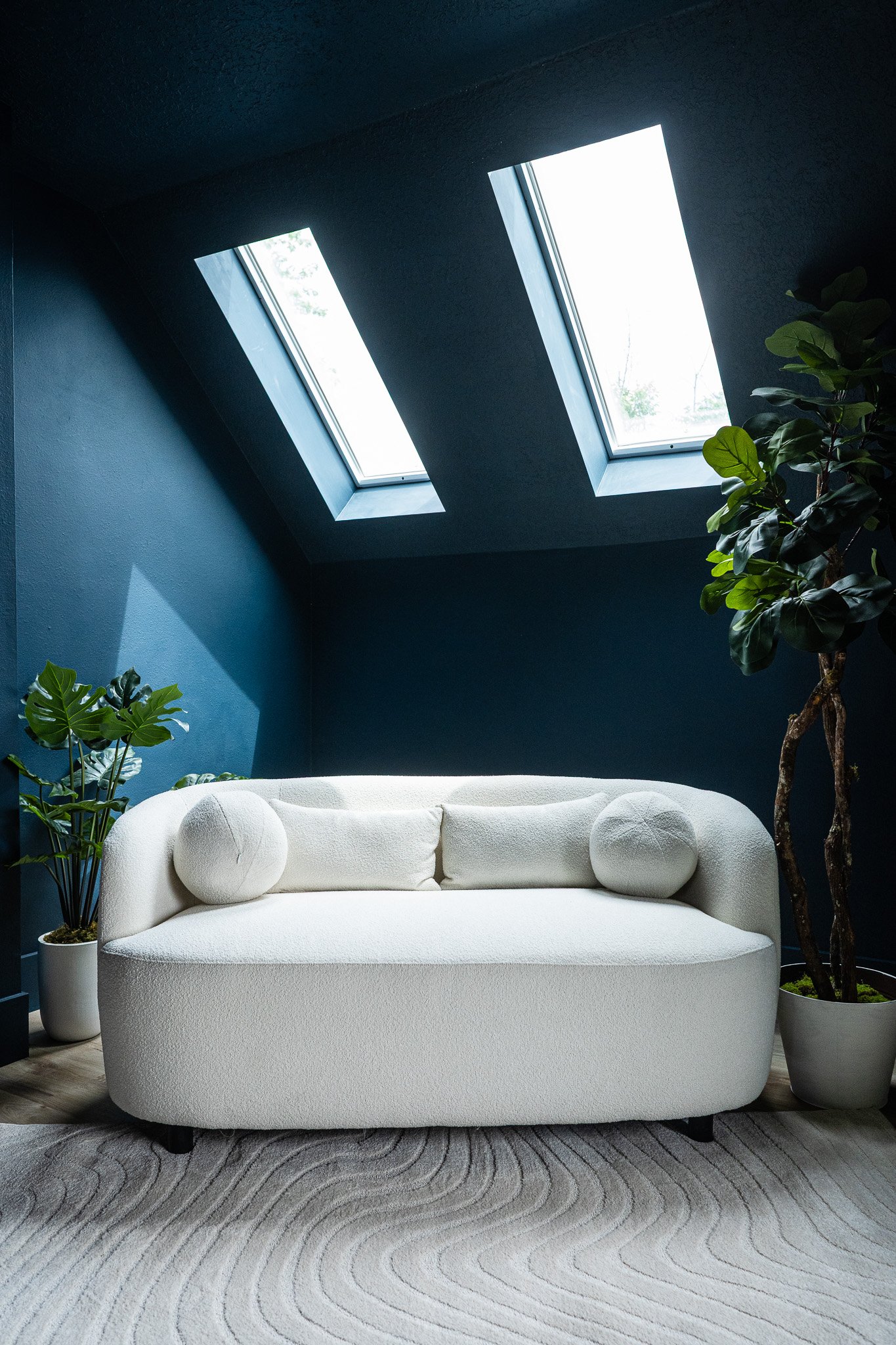 Modern sitting area with a white sofa, potted plants, and large skylights in a dark blue room.