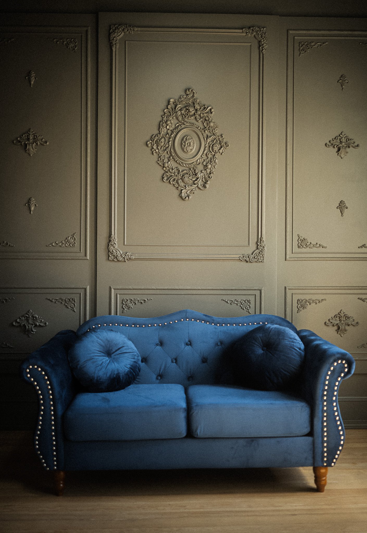 A vintage blue velvet sofa with button tufting and large round pillows on each side, set against a grey wall with ornate decorative molding and embellishments.