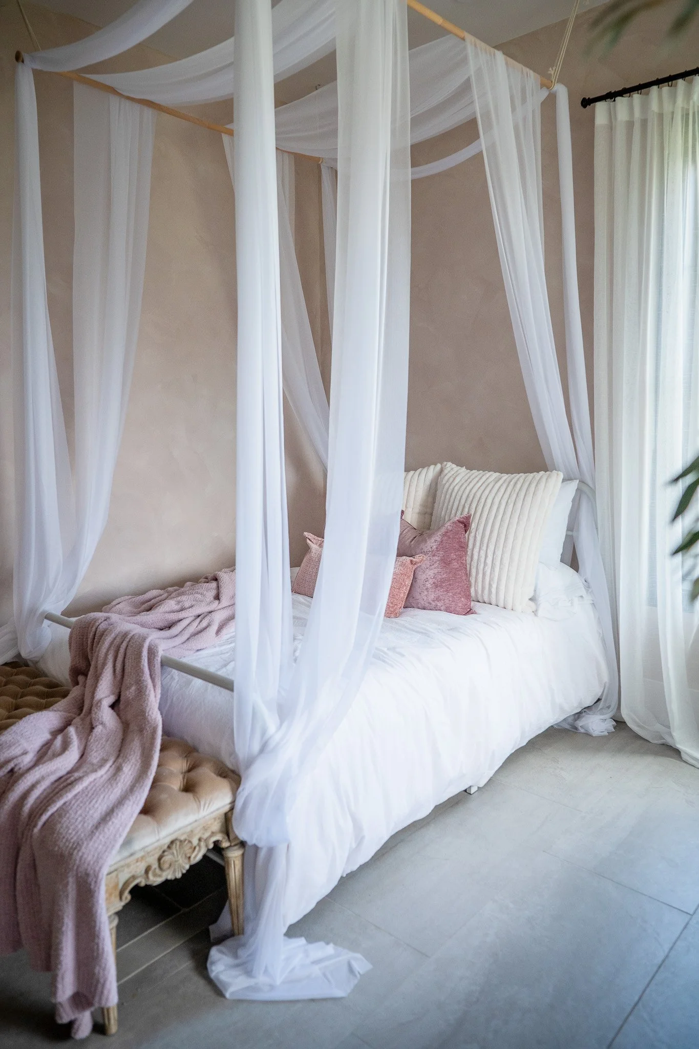 A luxurious bedroom with a canopy bed draped with white sheer curtains, decorated with pink and cream pillows, and a pink throw blanket on an ornate bench at the foot of the bed, next to a window with light curtains.