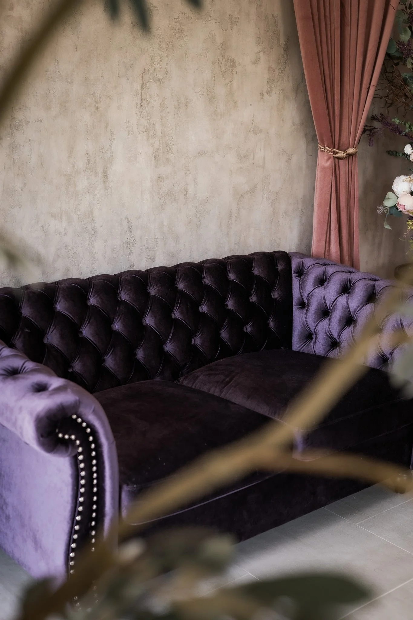A dark purple velvet tufted sofa with nailhead trim, placed against a beige textured wall, with a mauve curtain tied back on the right side.
