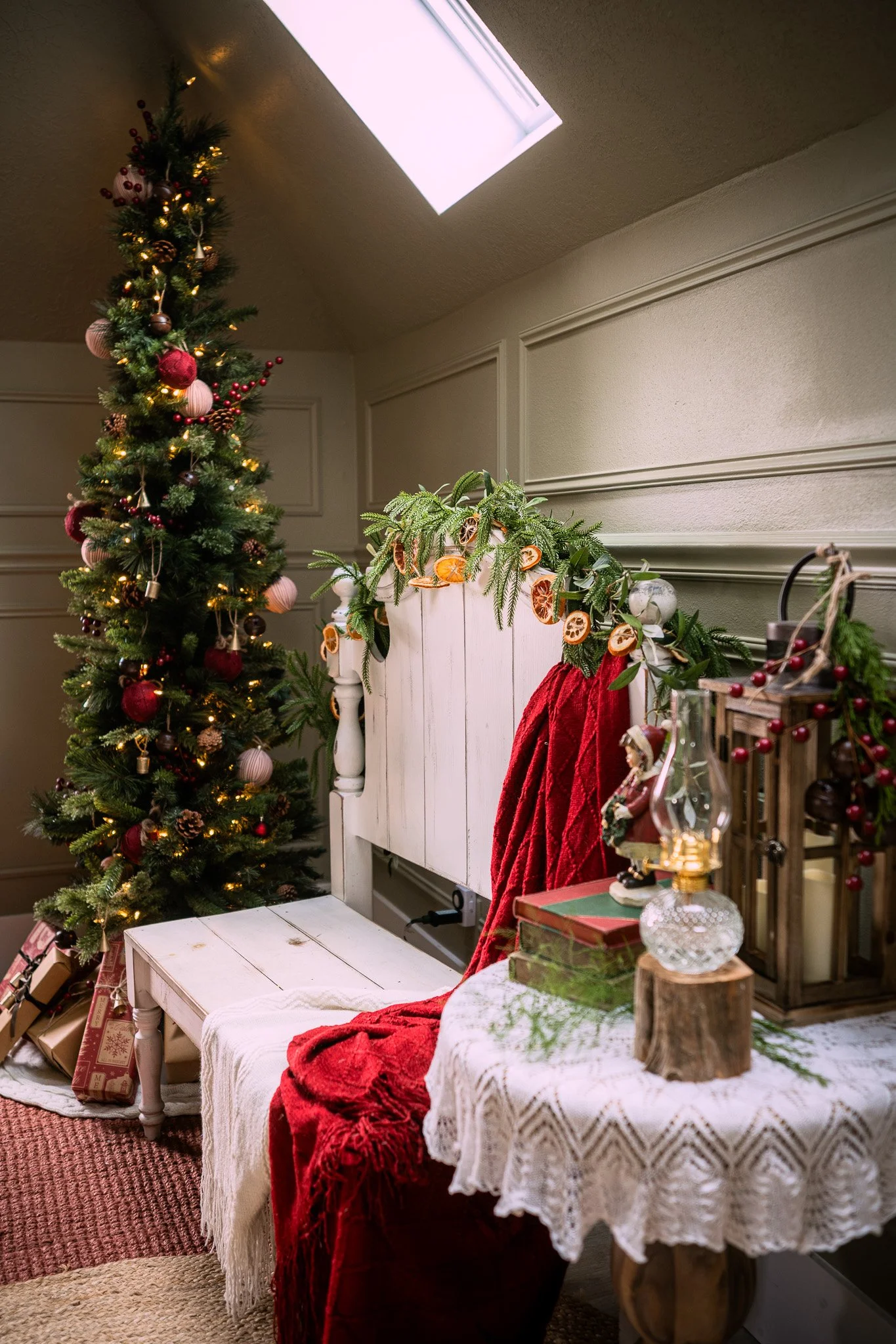 A decorated Christmas tree with lights and ornaments stands next to a white bench with a red throw blanket. Nearby, a small table with a lace tablecloth holds holiday decorations, including a lantern, candles, and a pine cone, with additional wrapped