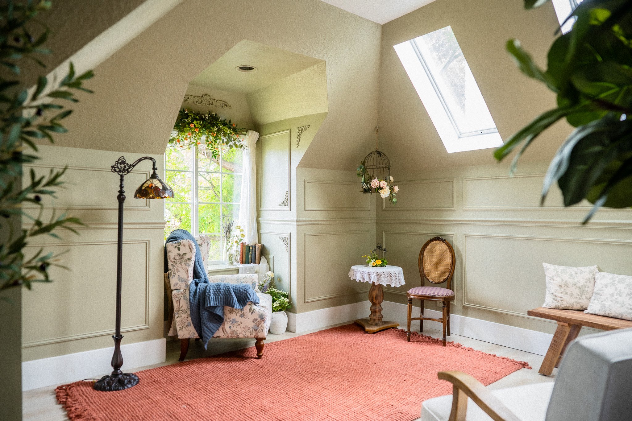 A cozy living space with vintage furniture, a floral armchair with a blue throw, a small round table with a yellow flower arrangement, a wooden bench with pillows, a tall floor lamp, and a framed window with curtains and greenery outside. The room ha