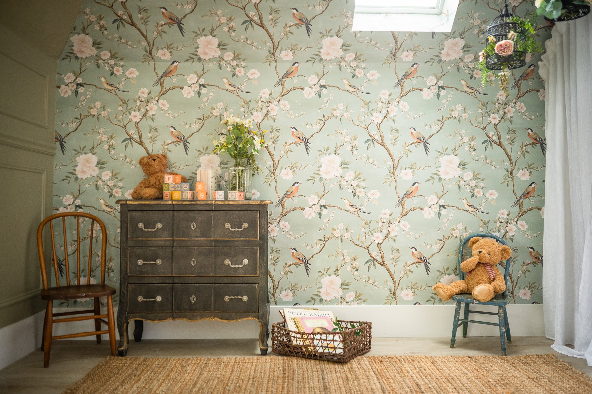 A cozy room with floral and bird-patterned wallpaper, a small wooden chair on the left, a vintage black dresser with a teddy bear, a vase with flowers, and colorful blocks on top. A wicker basket with books is on the floor, a blue chair with a teddy
