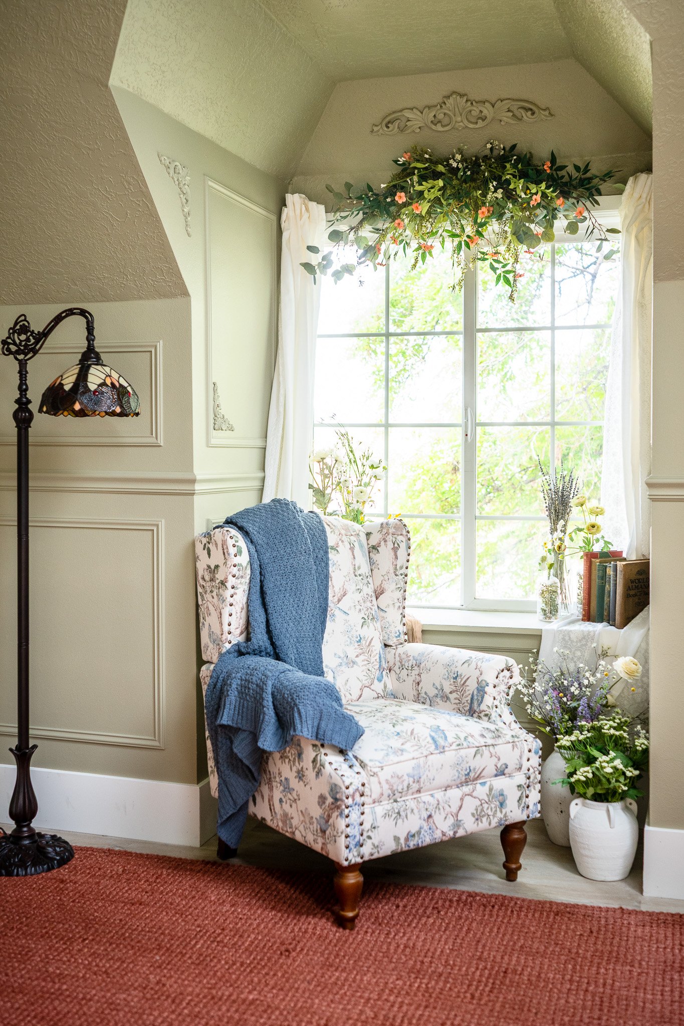 A cozy corner with a floral armchair draped with a blue blanket, surrounded by flowers in vases, a window with white curtains, and a hanging floral arrangement above.