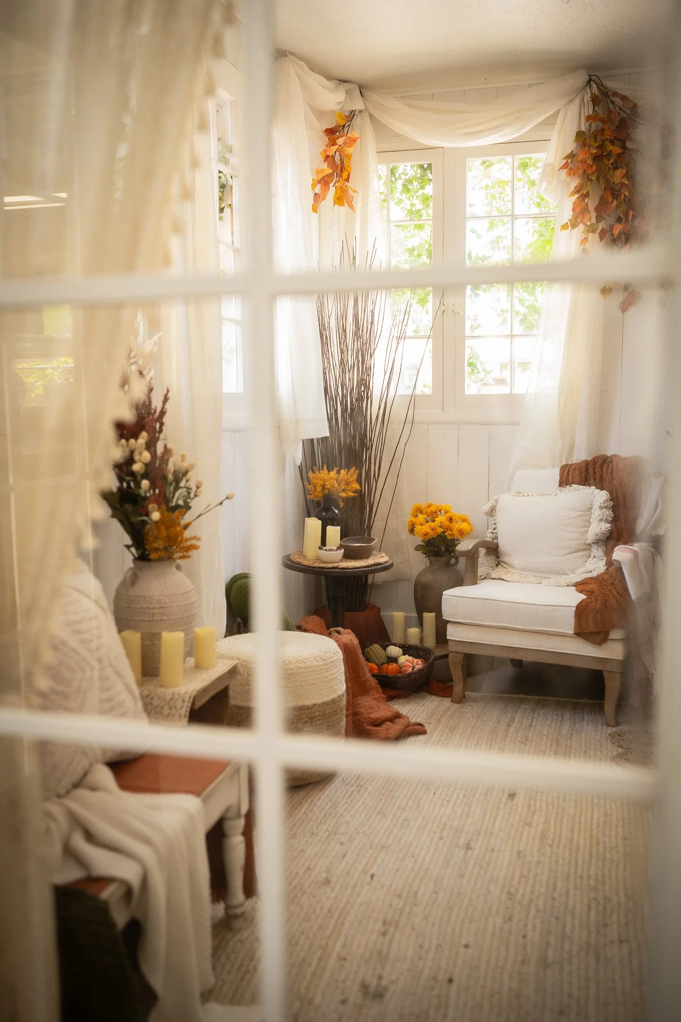 Cozy living room with white walls, large windows, white curtains, and autumn-themed decorations including orange and yellow flowers and fall leaves.