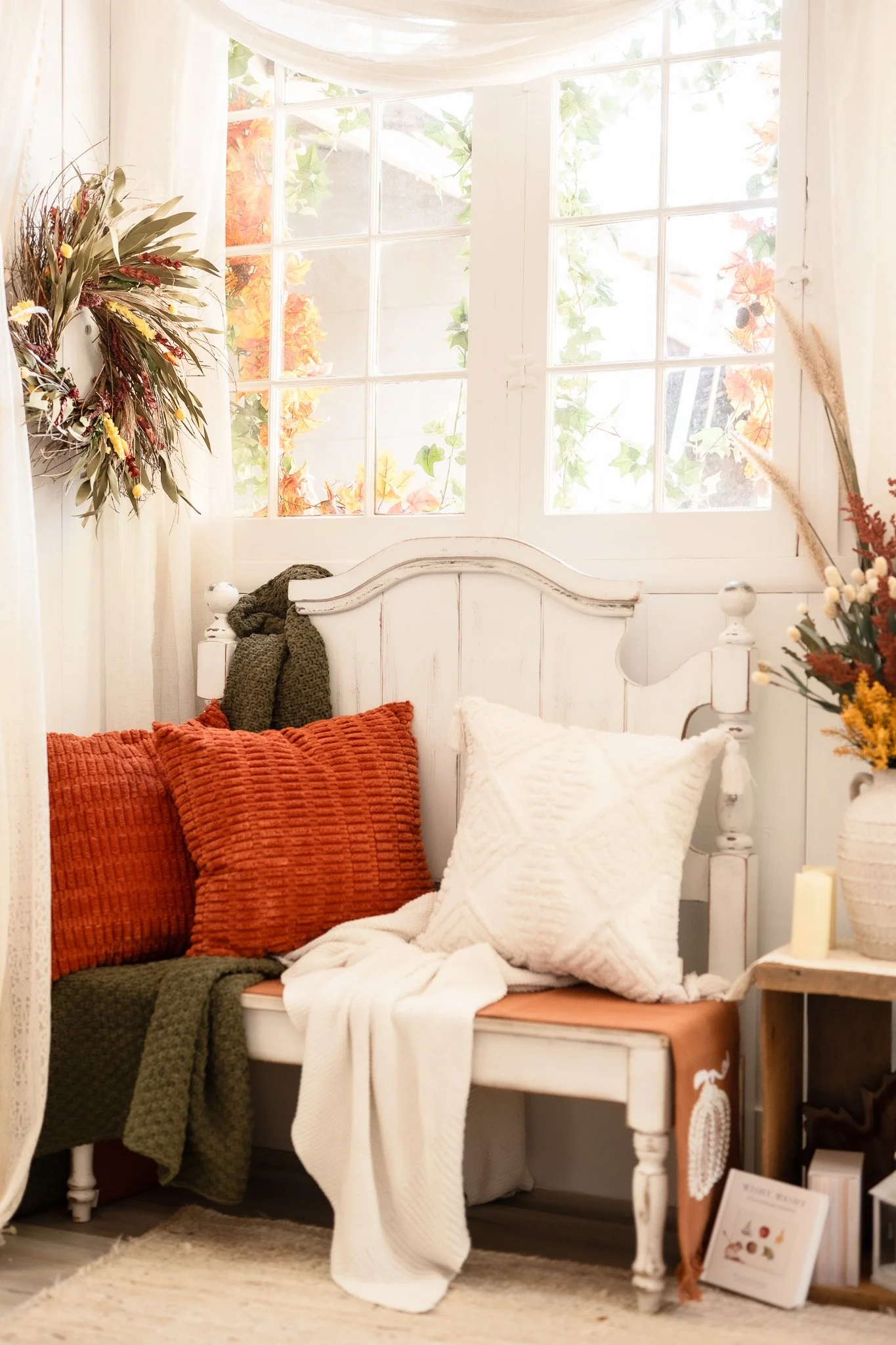 Cozy indoor corner with a white wooden bench decorated with textured orange and white pillows, a throw blanket, and surrounded by dried floral arrangements and a window with fall foliage outside.