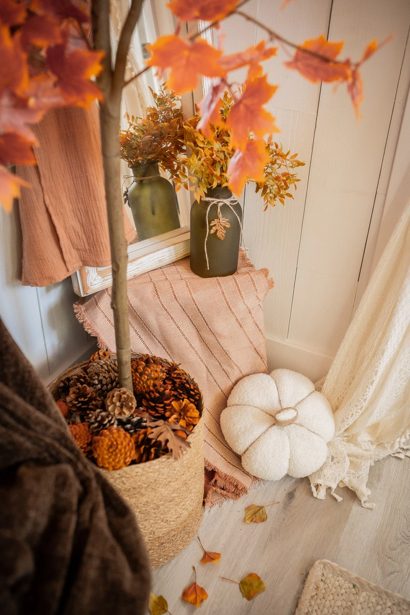 Decorative autumn-themed interior with a tree and basket of pinecones, a mirror, vases with fall leaves, a pumpkin-shaped cushion, and sheer curtains.