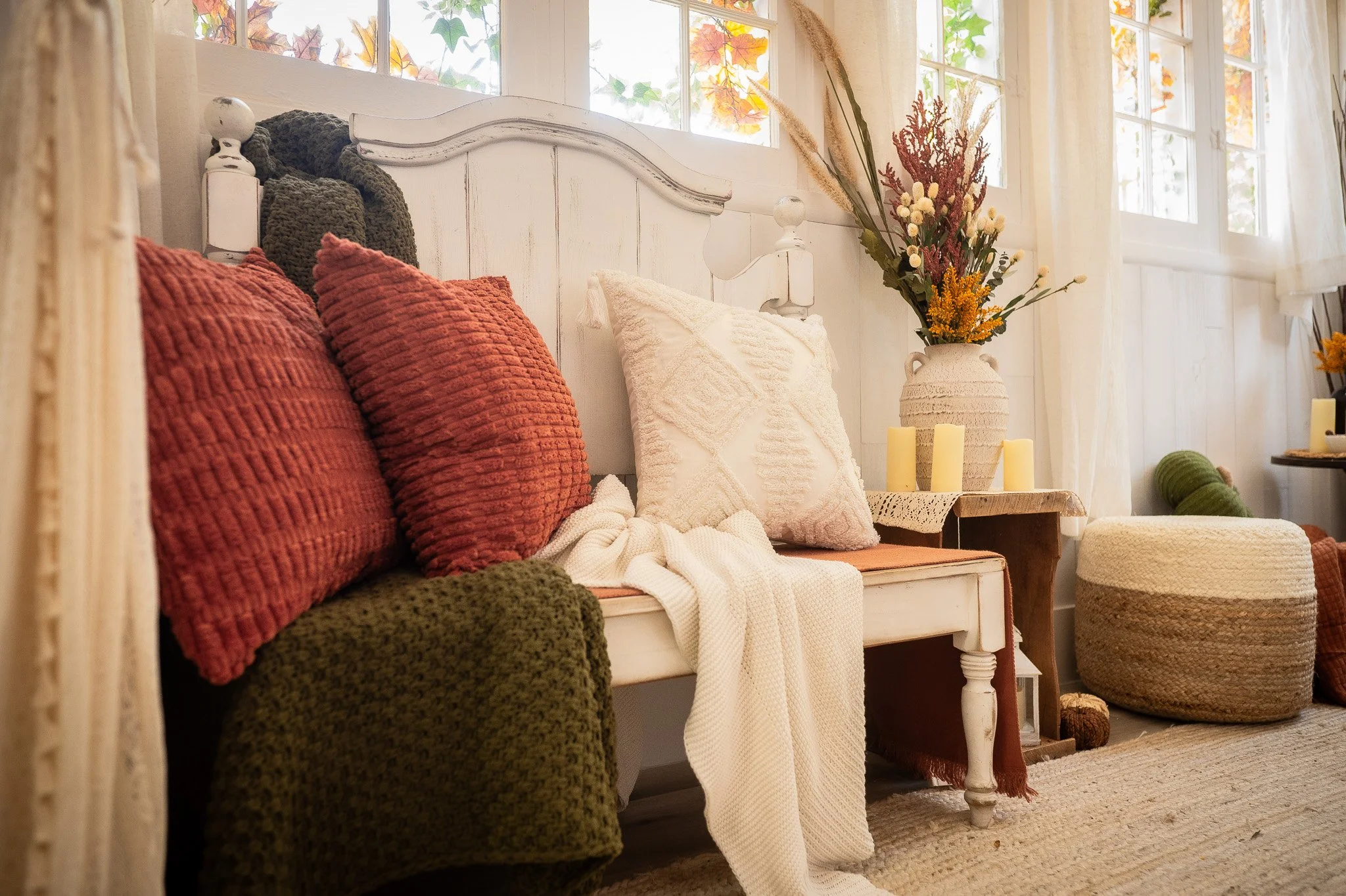 A cozy living room corner with a white wooden bench adorned with textured cushions in shades of red, green, and white. A knit throw blanket drapes over the bench. Next to it, a wooden side table holds a large vase filled with dried and fresh flowers,