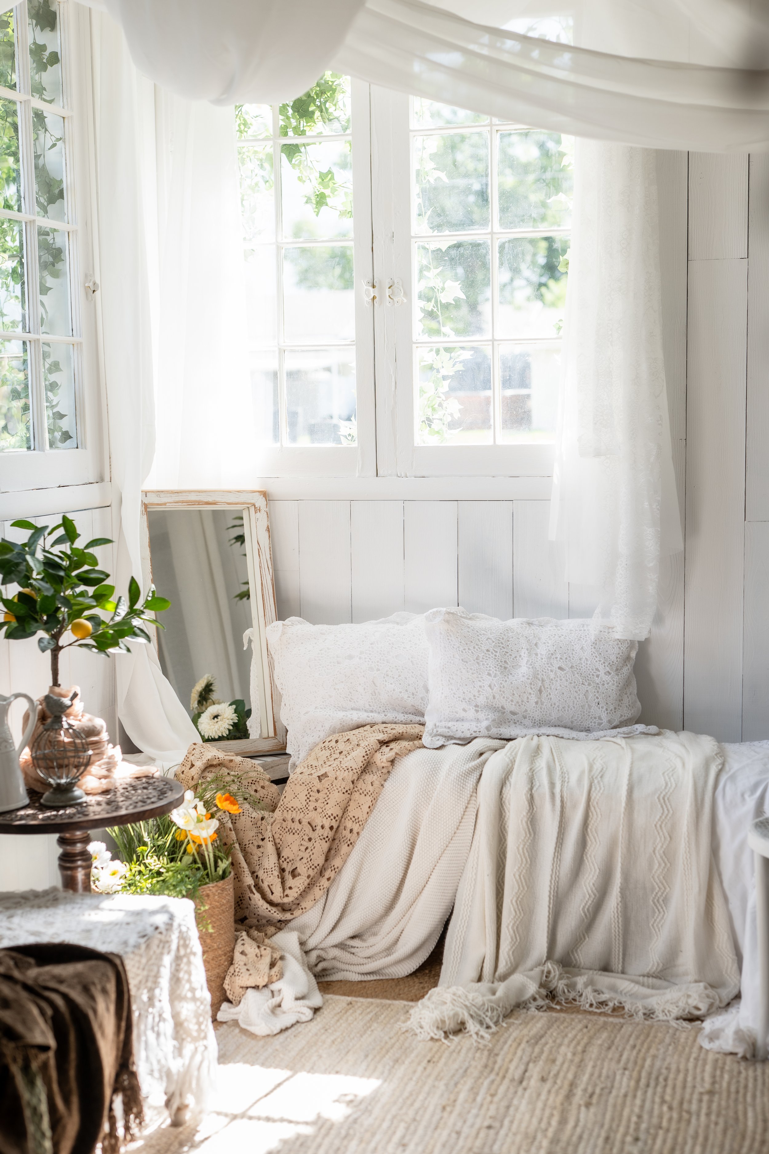 A cozy, sunlit corner of a room with white wood-paneled walls and a large window with sheer white curtains. There is a bed or seating area with white lace pillows and cream-colored blankets, a mirror with a distressed frame leaning against the wall,