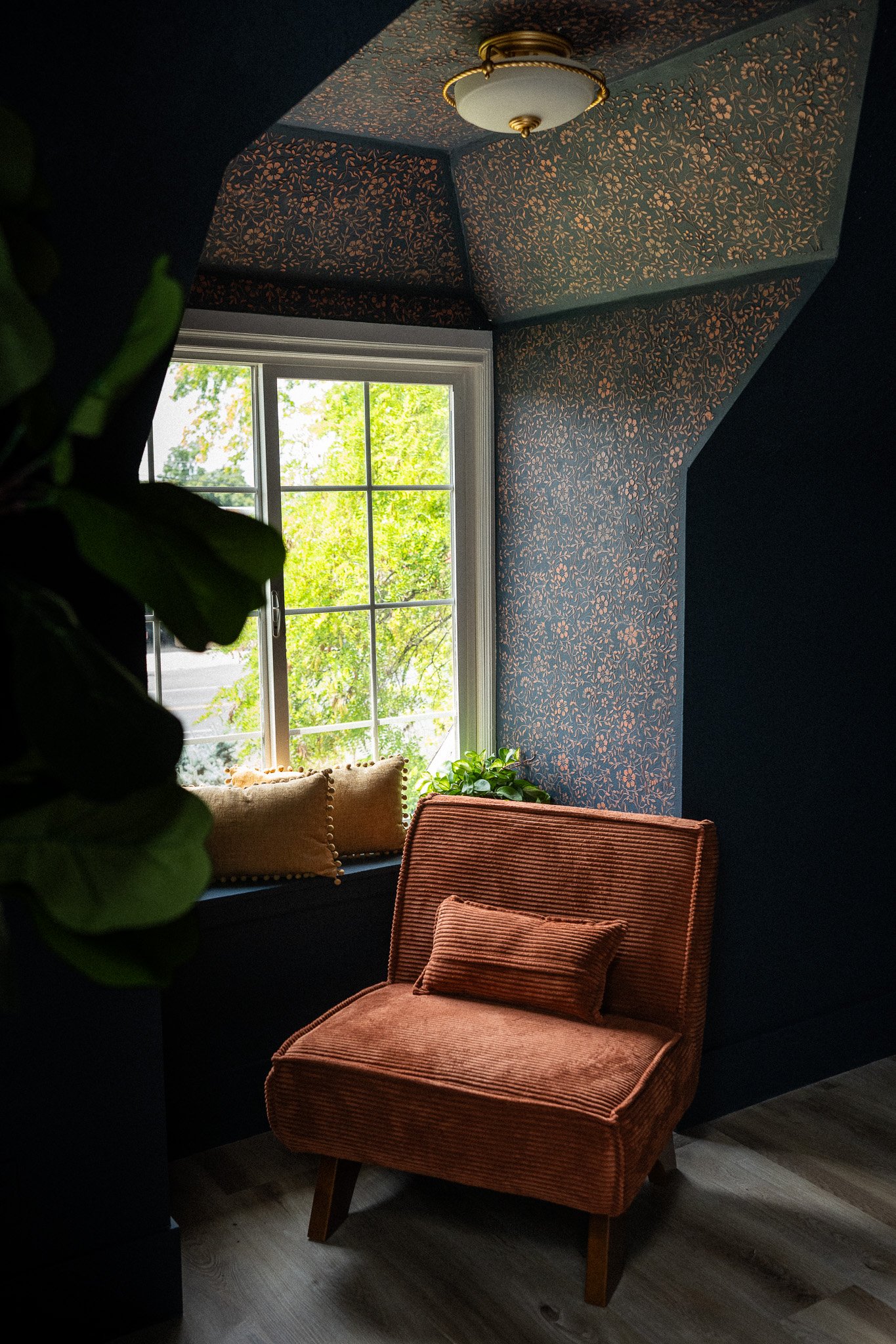 A cozy corner with a burnt orange corduroy chair, a window seat with pillows, and patterned wallpaper in a room with a ceiling light, greenery outside the window, and wooden flooring.