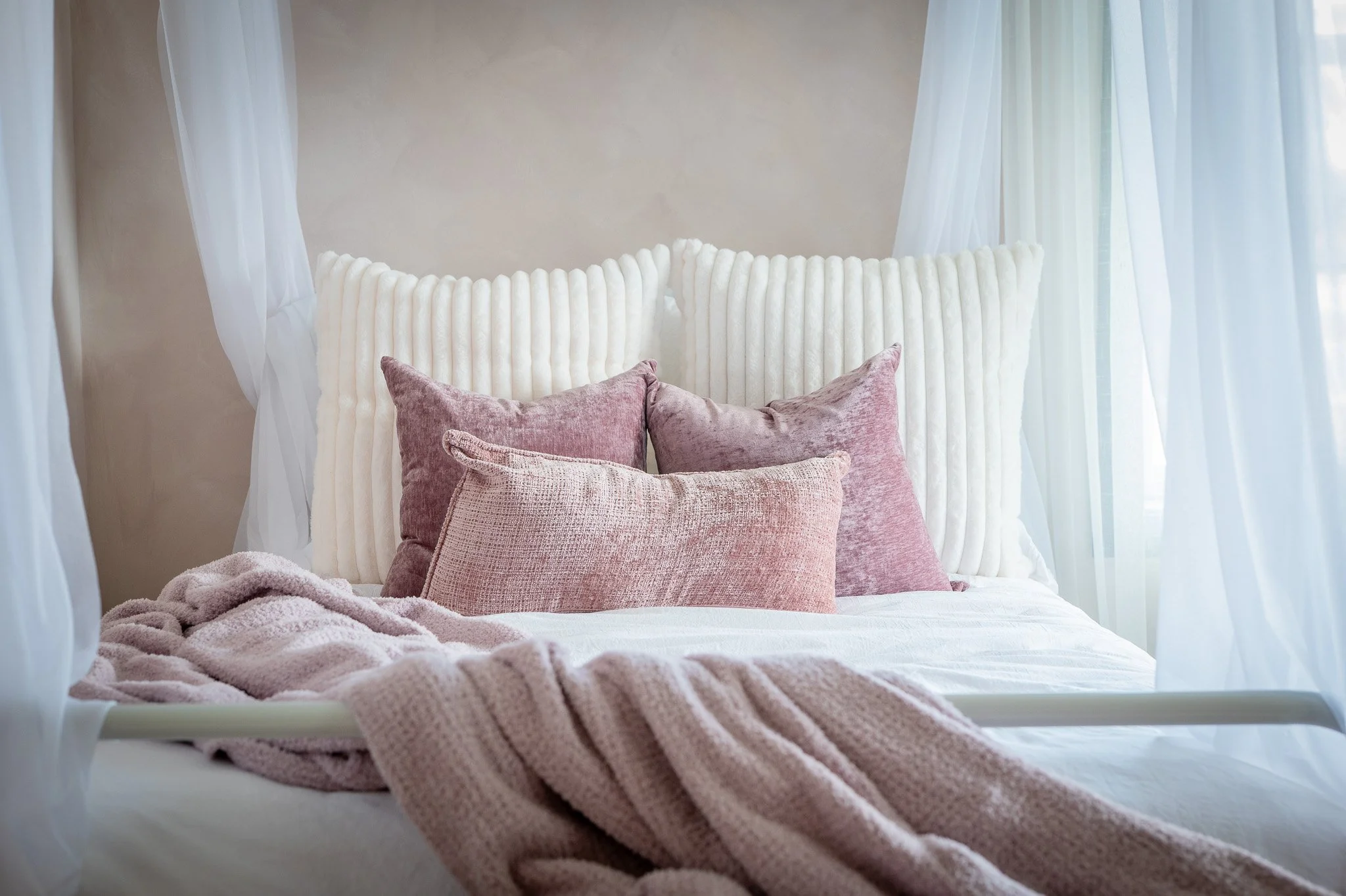 A neatly made bed with pink and white throw pillows, a pink blanket, and white bed linens, framed by sheer white curtains in a softly lit bedroom.