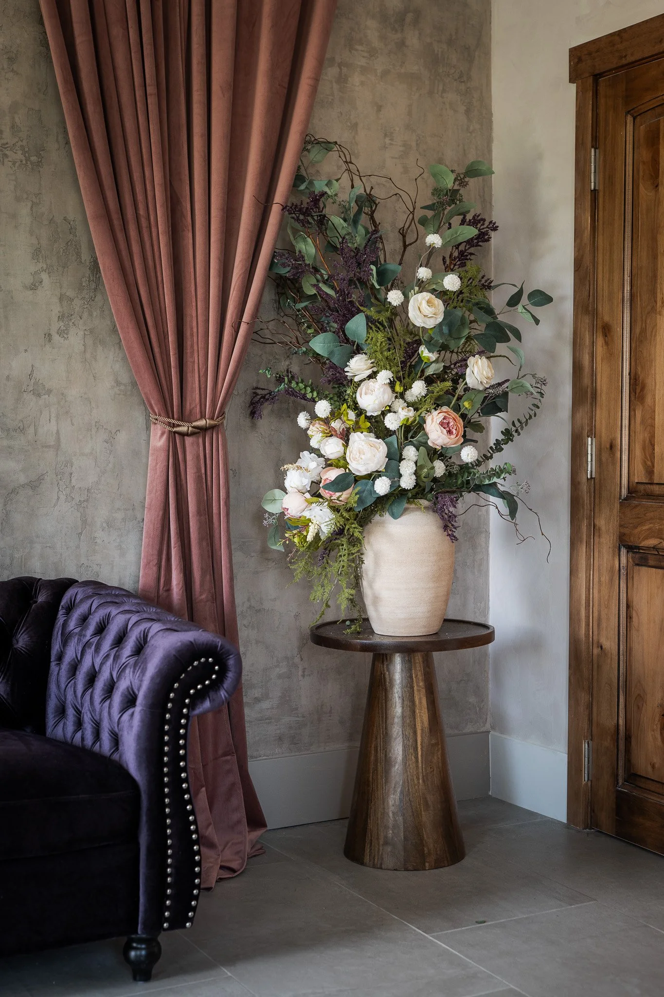 A large floral arrangement with white and pink flowers, green leaves, and branches in a cream-colored vase on a wooden table next to a deep purple tufted sofa and a pink curtain in a room with concrete walls and wooden door.