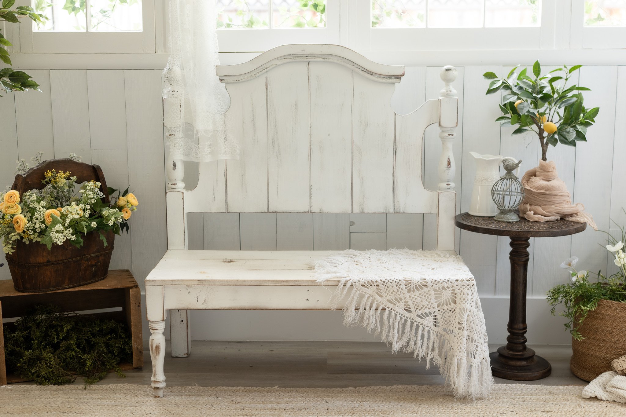 A vintage white wooden bench with a lace throw, flanked by a flower basket on a small wooden stand on the left and a small round table with decorative items and a potted lemon tree on the right, in a bright room with white paneled walls and a window.