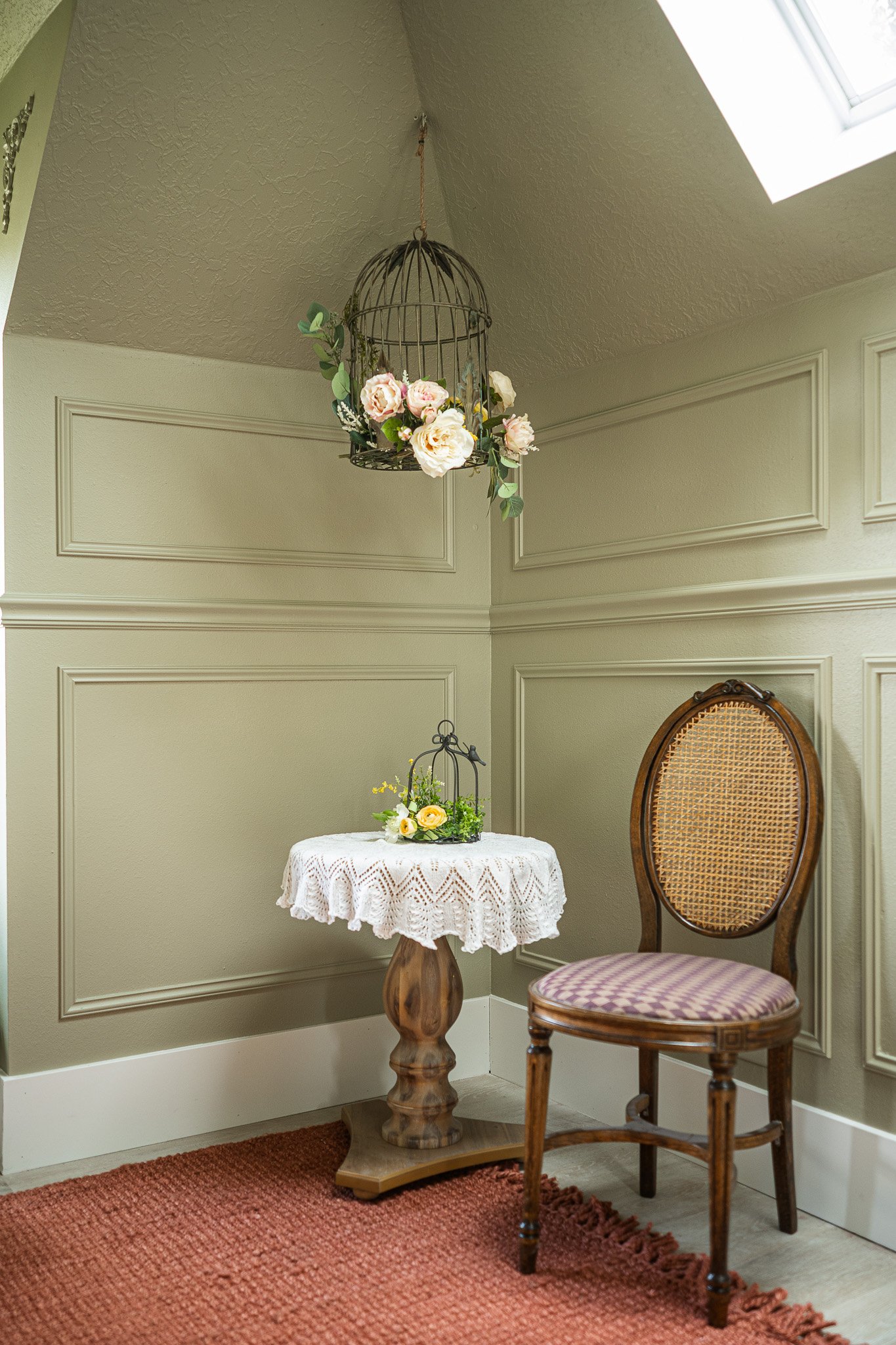 A corner of a room with light green walls, a wooden chair with a woven back and cushioned seat, a small wooden table with a white lace tablecloth, and decorative birdcages with flowers hanging and resting on the table. A skylight illuminates the scen