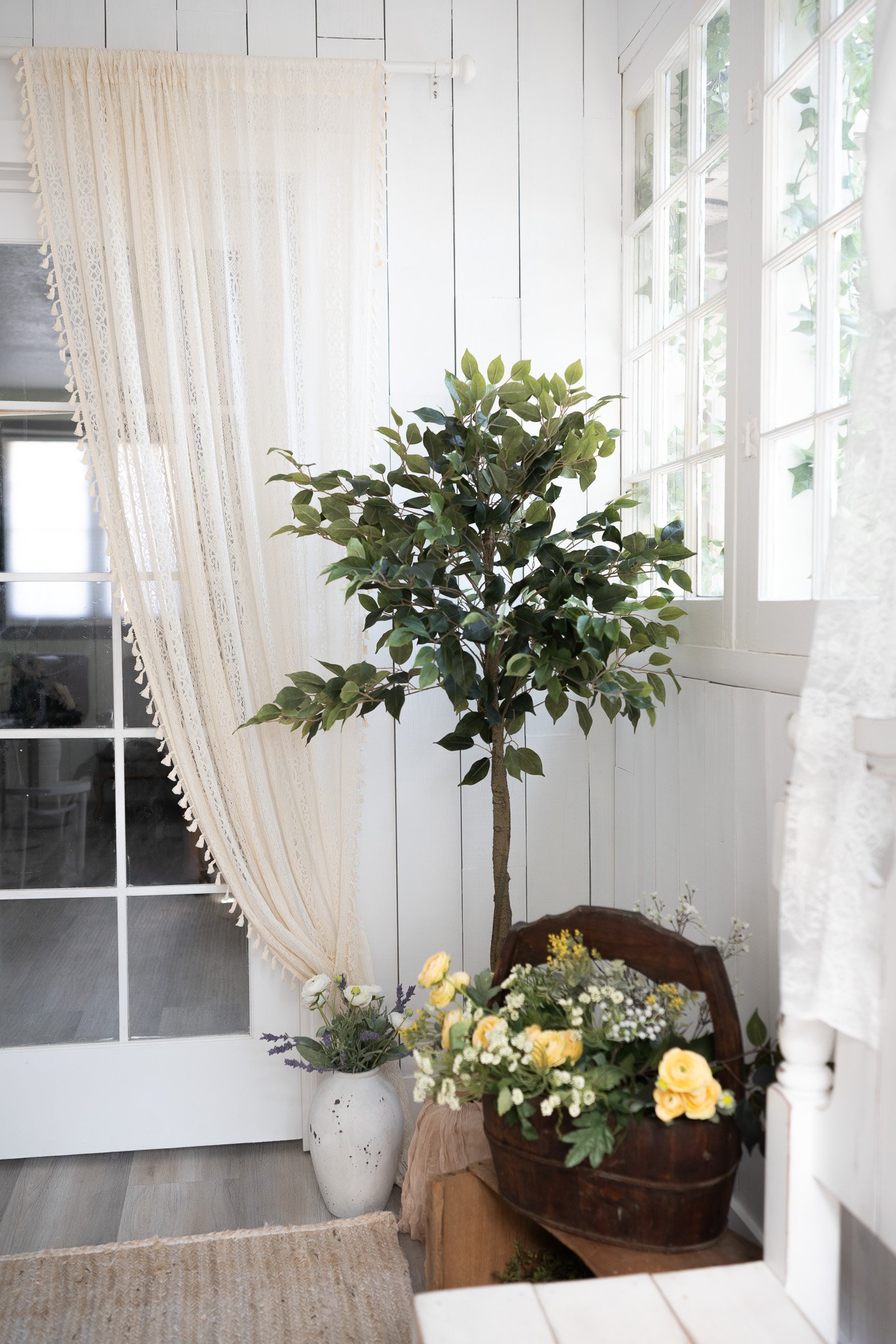 Indoor corner with a large potted artificial tree, a wooden chest decorated with a flower arrangement, a white vase with flowers, lace curtains, and a window with natural light.