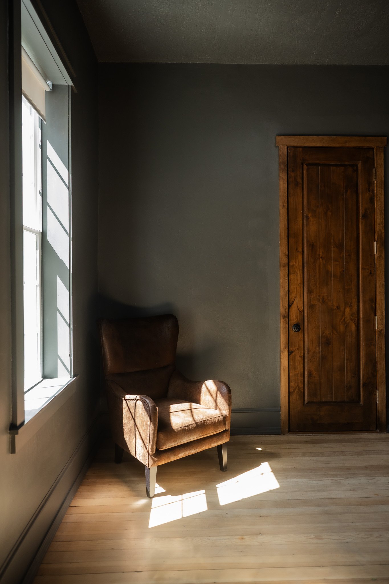 Sunlight streams through a window onto a brown armchair in a room with dark walls and a wooden door.