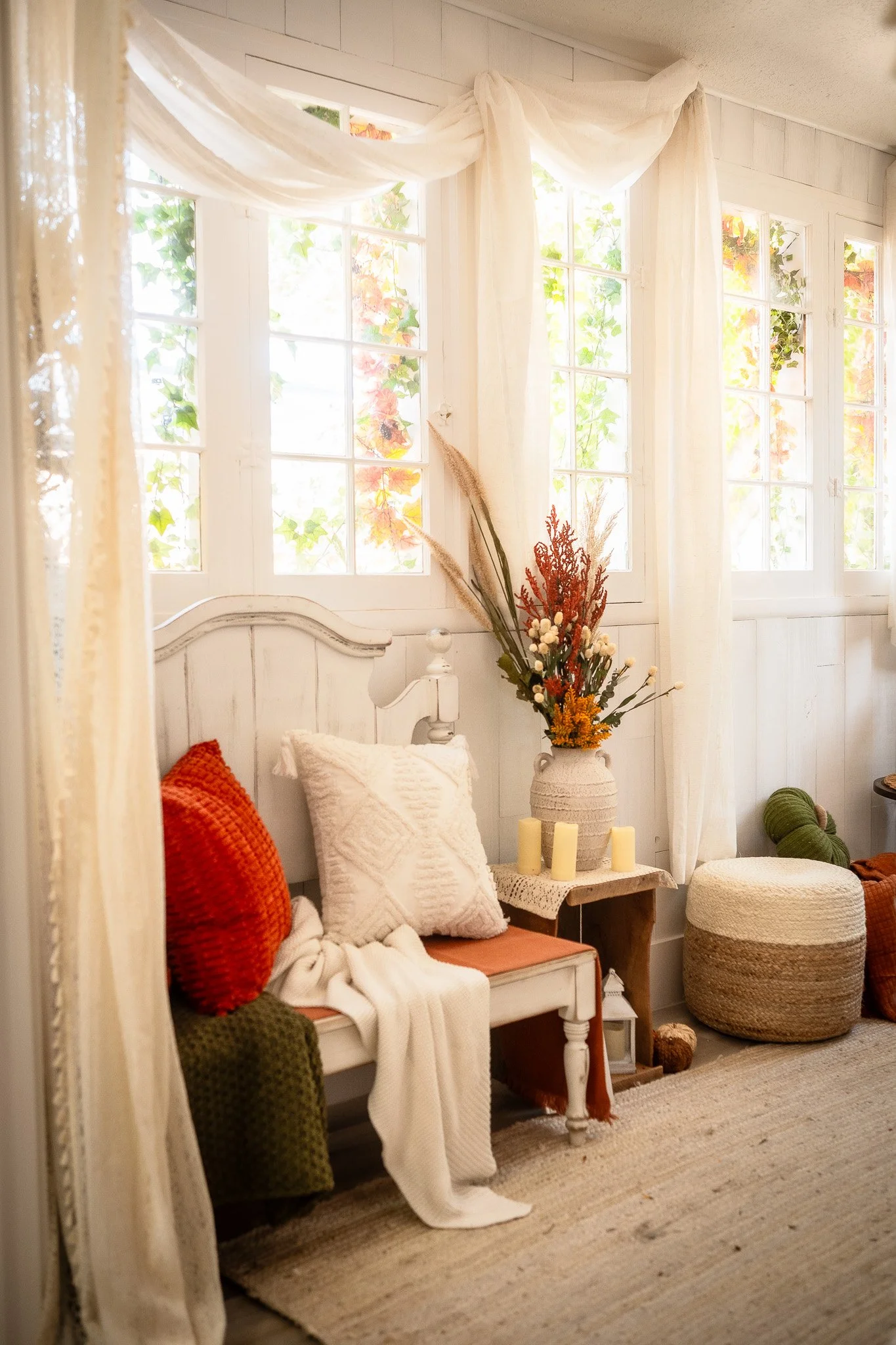 A cozy interior room with white wooden walls and large windows with cream curtains. There's a white bench with orange and white pillows, a beige throw blanket, and a small side table with a woven vase of dried flowers and three pillar candles. A knit