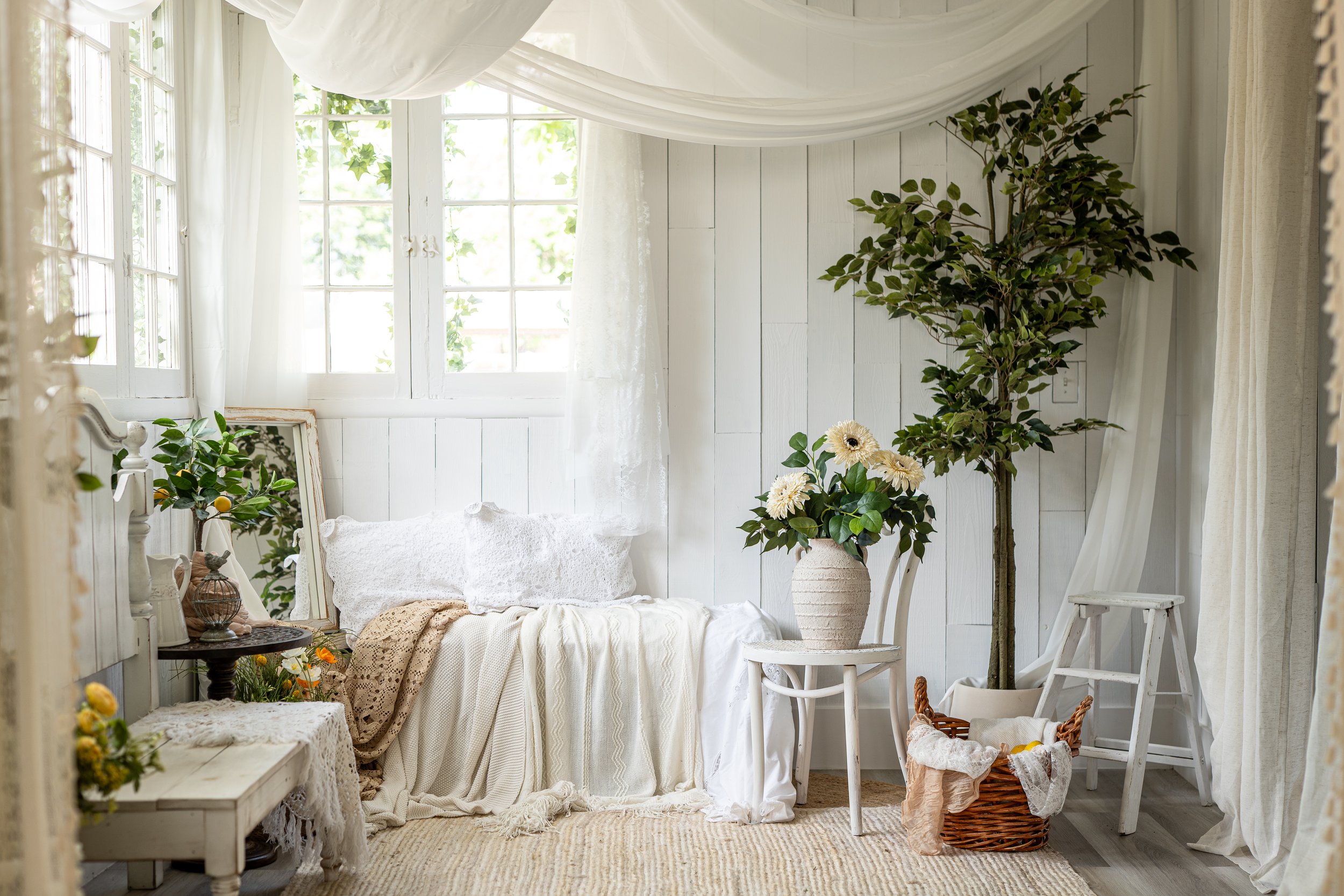 Bright, cozy room with white wooden walls, windows with sheer curtains, and plants, including a tall tree and potted flowers, with white and neutral-colored furniture and decor, creating a serene atmosphere.