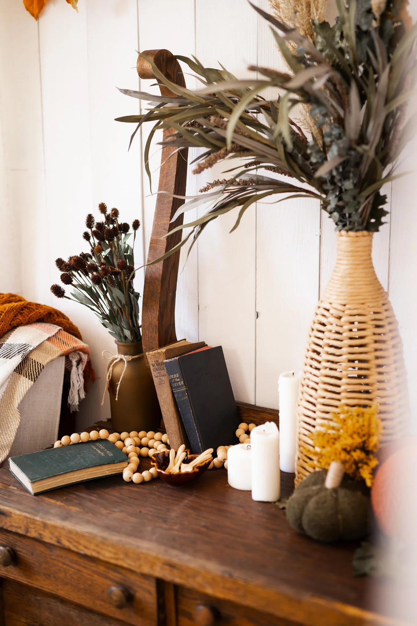Decorative arrangement on a wooden dresser includes a tall woven vase with dried flowers, a smaller brown vase with dried flowers, a stack of books, candles, a string of wooden beads, and a small pumpkin. Part of a plaid blanket is visible on the lef