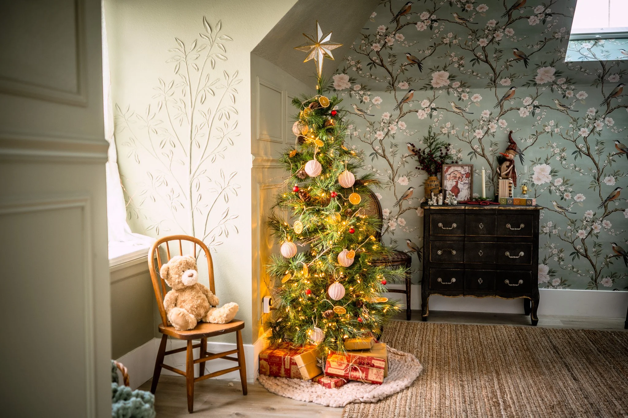 A decorated Christmas tree with lights, ornaments, and a star topper sits in a cozy living room corner, with wrapped presents underneath. To the left, a wooden chair holds a teddy bear, and to the right, a black dresser with decorative items is again