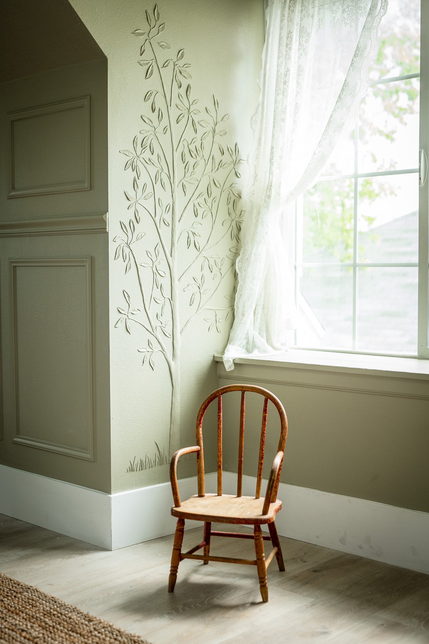 A small wooden chair in front of a white wall with a painted tree design, next to a window with sheer lace curtains letting in natural light.
