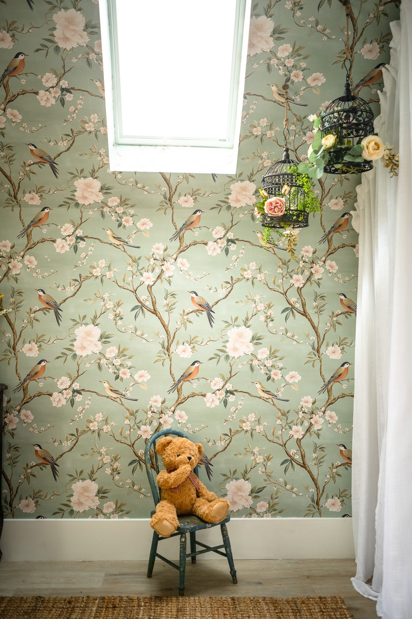 A room with floral wallpaper featuring birds, a small blue chair with a teddy bear, and hanging birdcage decor with flowers near a window.