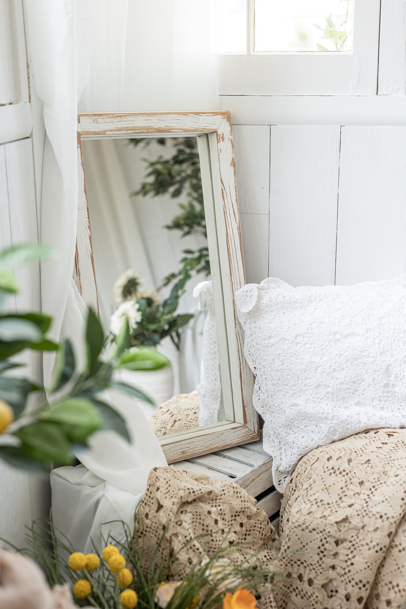 A mirror resting against a white wall, surrounded by a white lace pillow, a crochet blanket, and a bouquet of yellow flowers in a white vase.