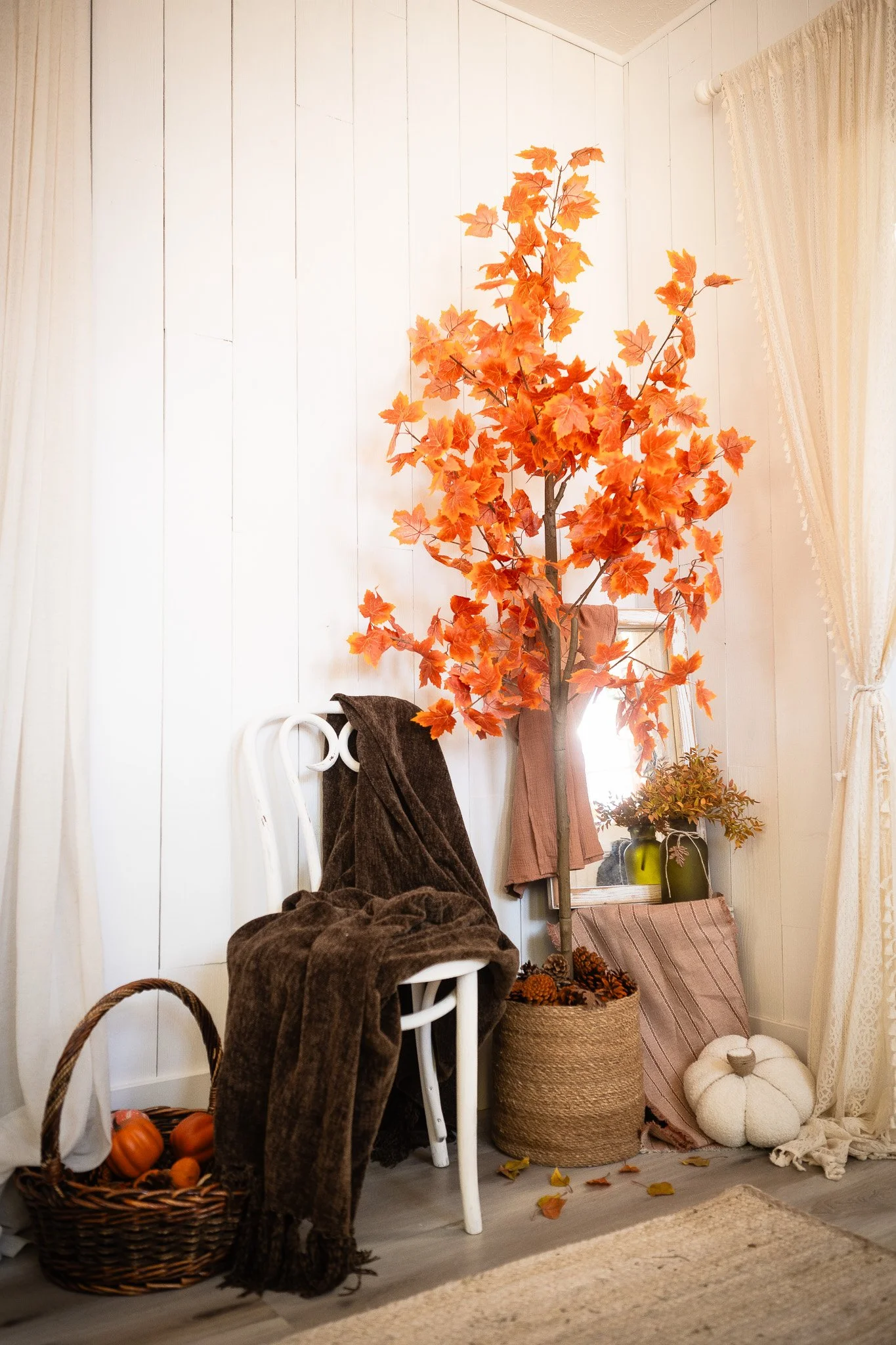 Decorative fall-themed corner with a white chair covered in a brown blanket, a basket of mini pumpkins, an autumn-colored artificial tree, a mirror, and fall decor accessories.
