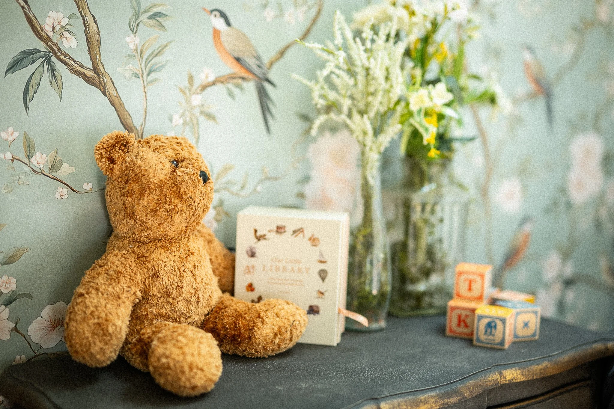 A teddy bear sits on a dark surface with a floral and bird-patterned wallpaper in the background. There are yellow and white flowers in glass vases, a small book, and colorful alphabet blocks on the surface.