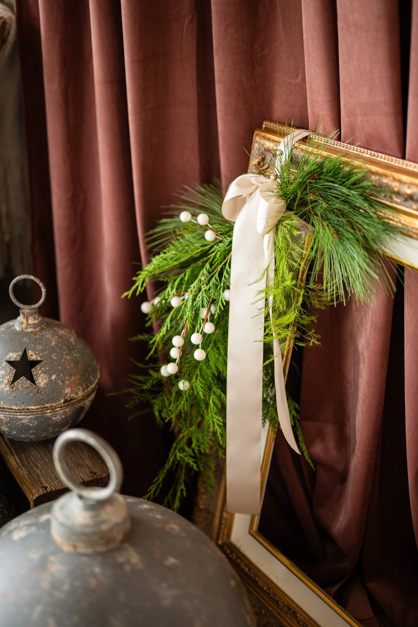Decorative Christmas wreath with white ribbon and green pine branches, placed on an ornate gold frame against mauve curtains, with rusty metal lanterns nearby.