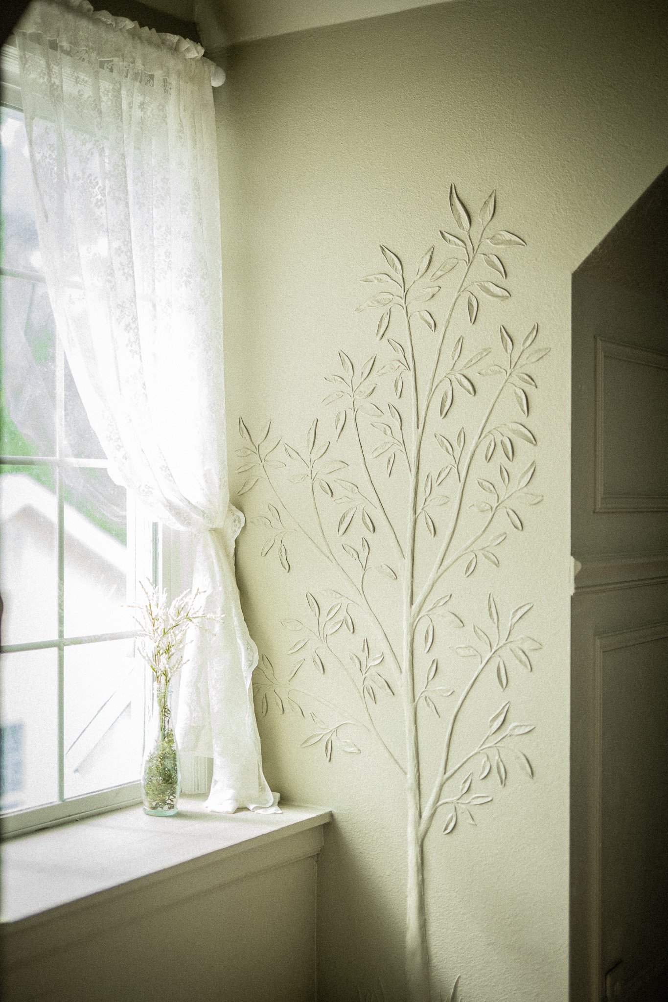 Sunlight coming through a lace curtain on a window with a decorative wall art of a tall plant or tree with multiple branches and leaves on a pale yellow wall, and a small vase with dried flowers on the window ledge.