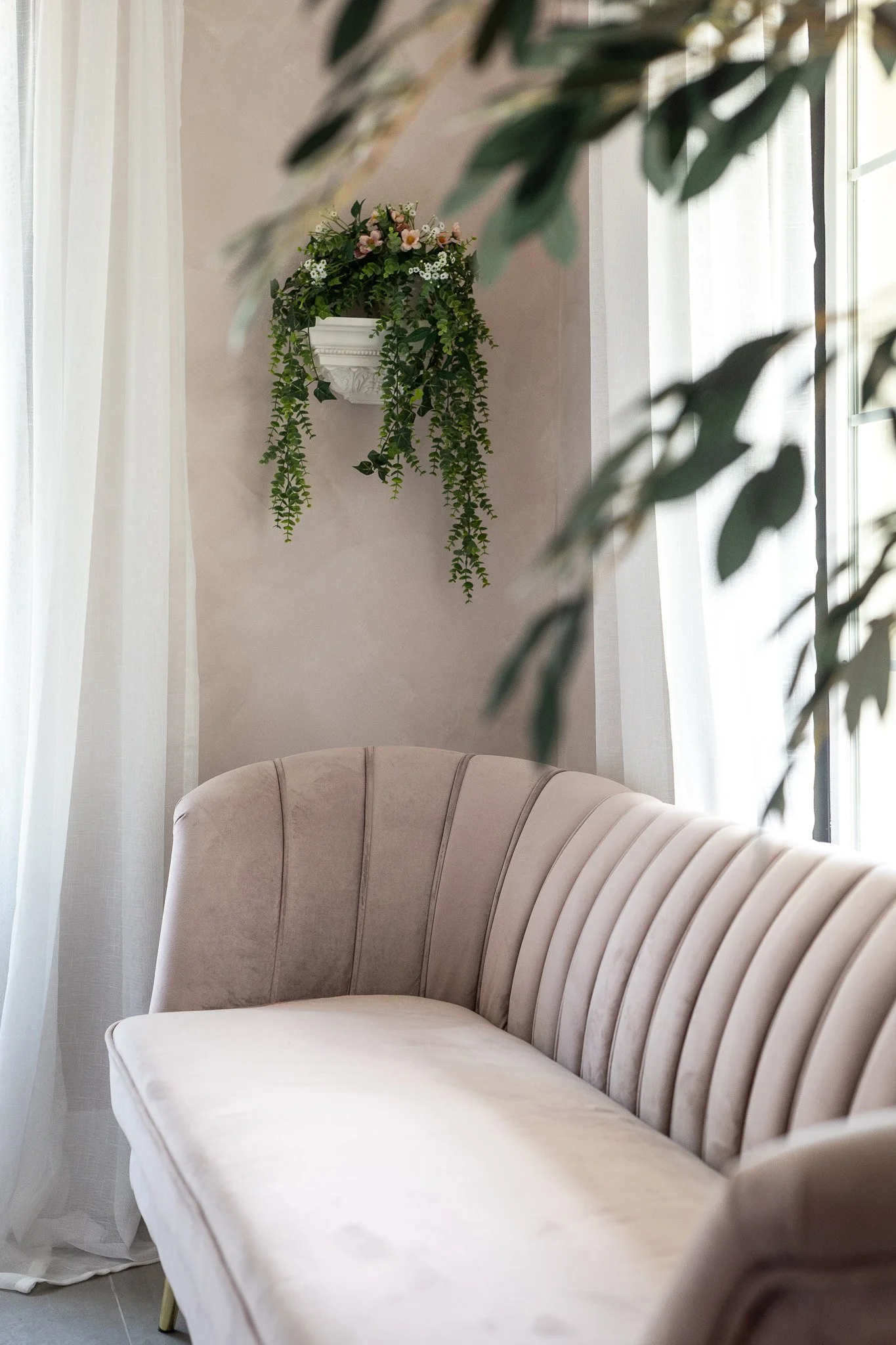 A beige velvet sofa with vertical stitching, next to a large window with white curtains, with a wall-mounted white flower planter filled with pink and white flowers and trailing green vines.
