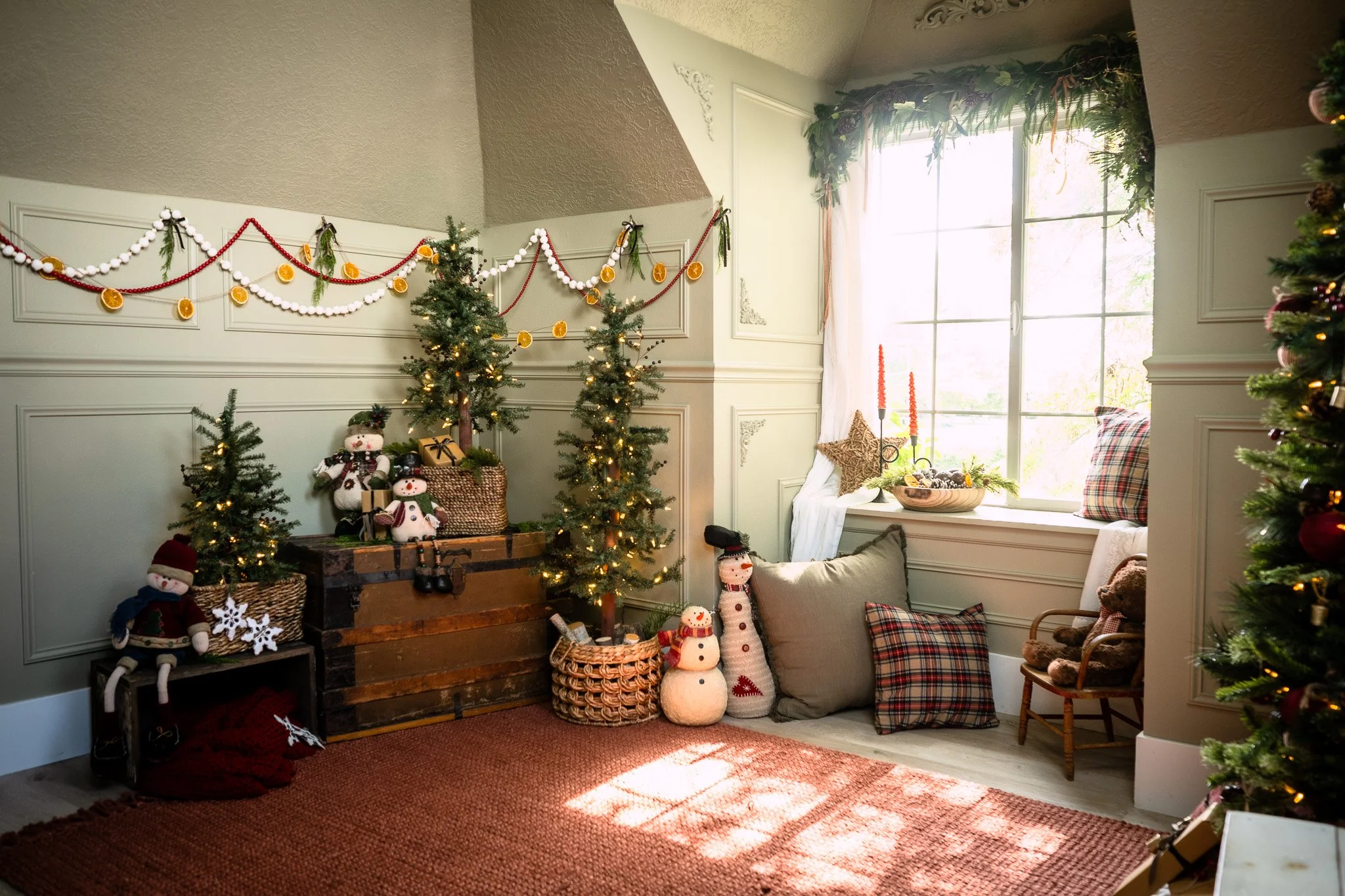 A cozy holiday living room decorated for Christmas with small Christmas trees, plush snowmen, and fairy lights, near a large window with natural light.