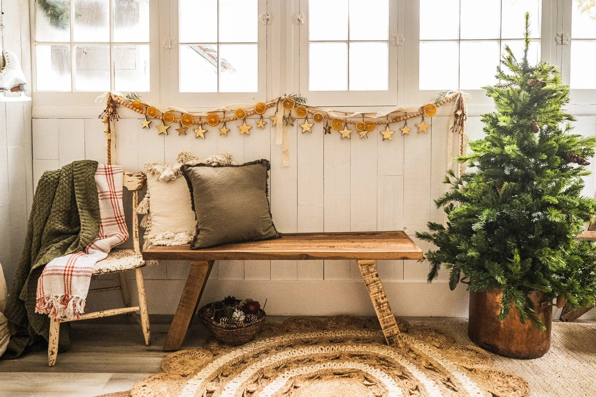 Decorative indoor space with a Christmas tree, pillows on a chair and bench, holiday garland on the wall, and a basket of ornaments on the floor.