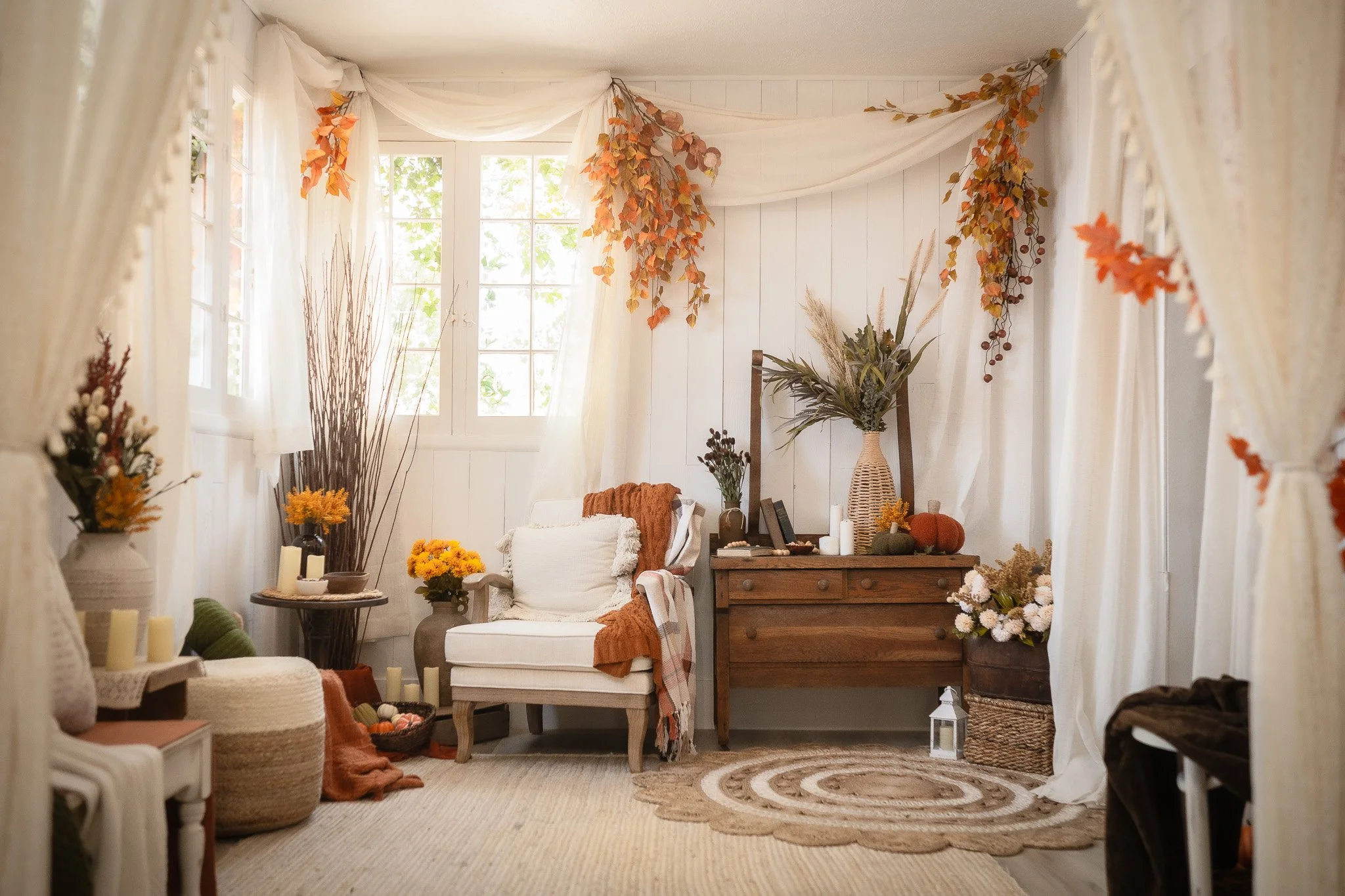 Cozy bohemian-style living room with white walls, cream curtains, autumn-colored leaves hanging from the ceiling, and a window letting in natural light. Decor includes dried flowers, pumpkins, candles, woven baskets, and a wooden dresser with plants