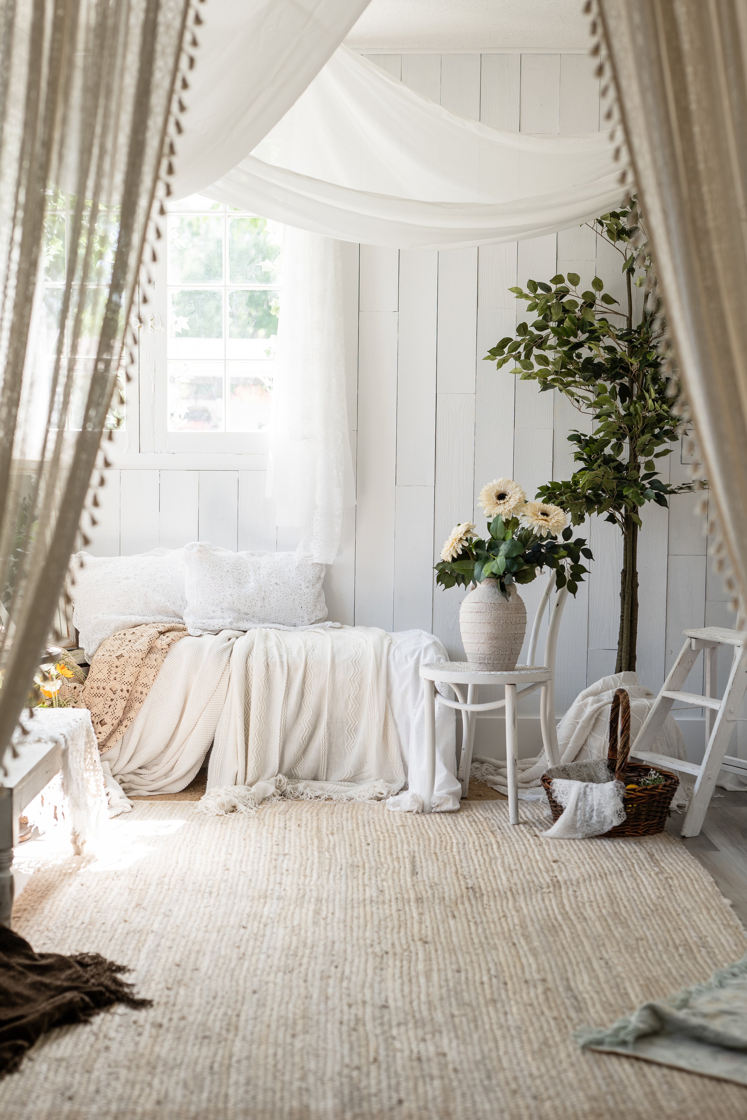 Bright bedroom with white paneled walls, sheer white curtains, a window with natural light, a bed with white bedding, a white chair with a large vase of white flowers, a tall green plant, a woven basket, and a beige jute rug.