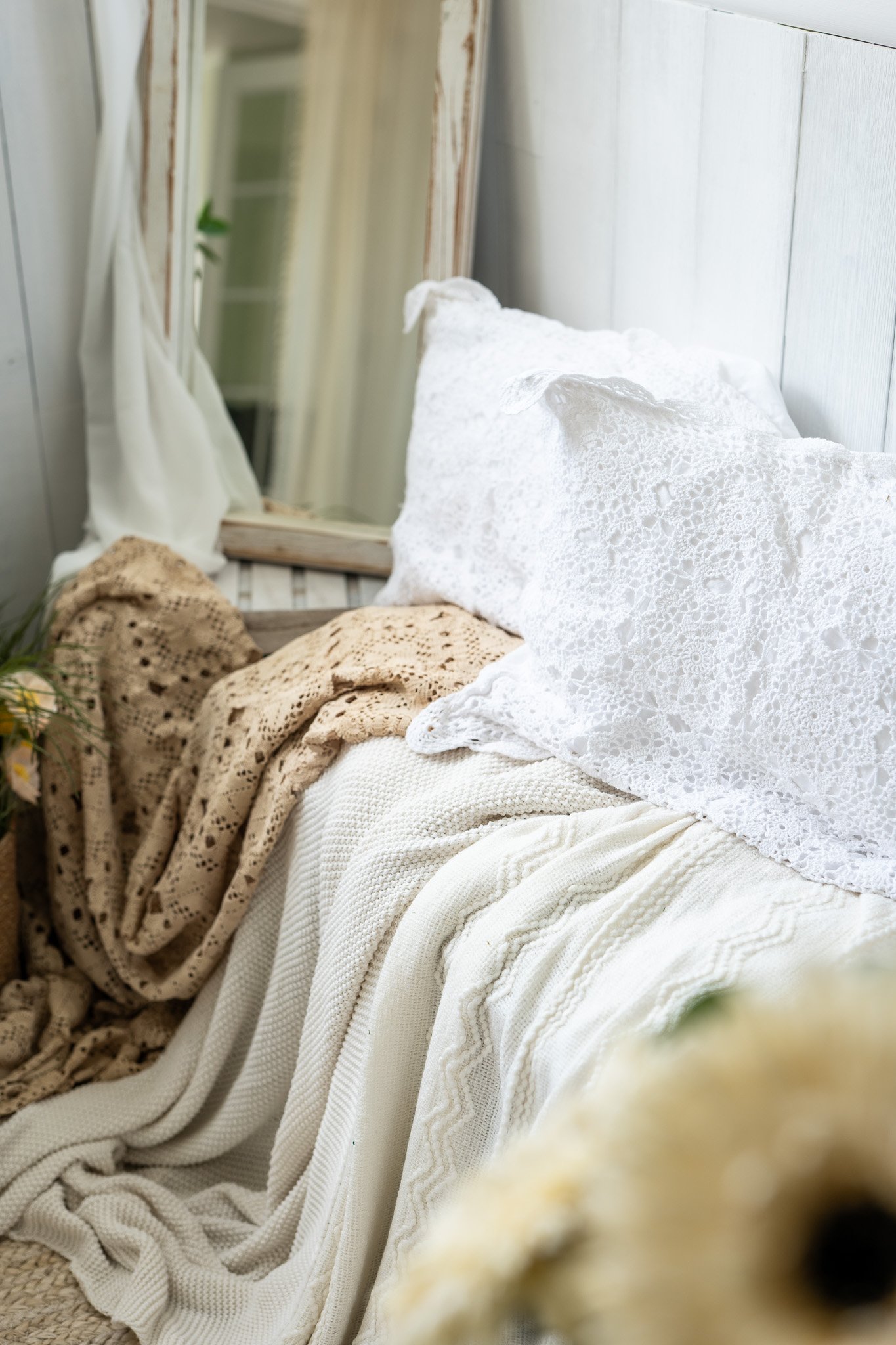 A cozy, vintage-style sofa with white lace and beige knitted blankets, and white lace pillows, with a mirror and white wall in the background.