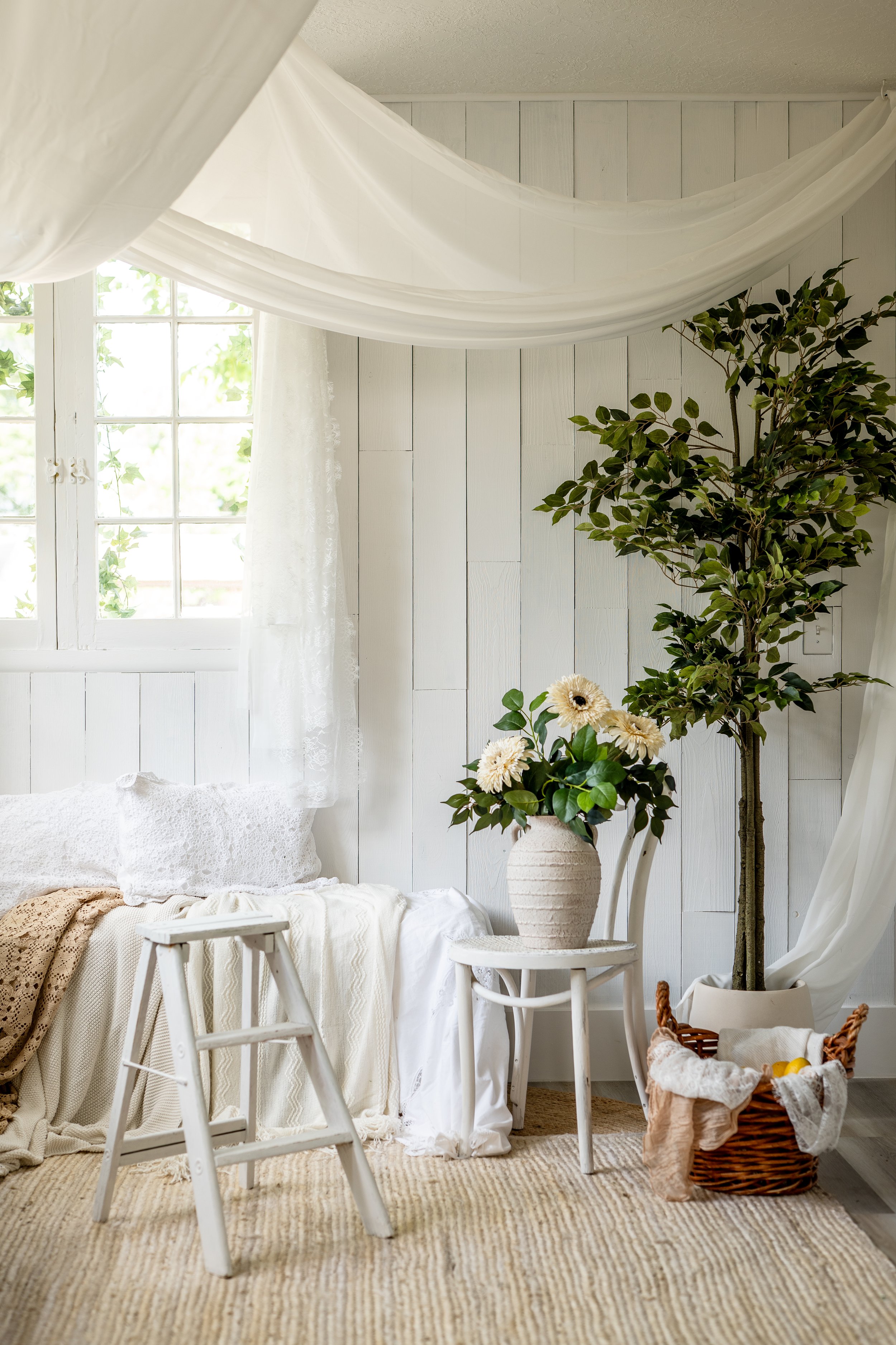 Interior of a bright room with white wooden walls, a window with lace curtains, a bed with white bedding, a small white table with a vase of flowers, a plant in a white pot, and a wicker basket with towels and lemons, all in a neutral, rustic style.
