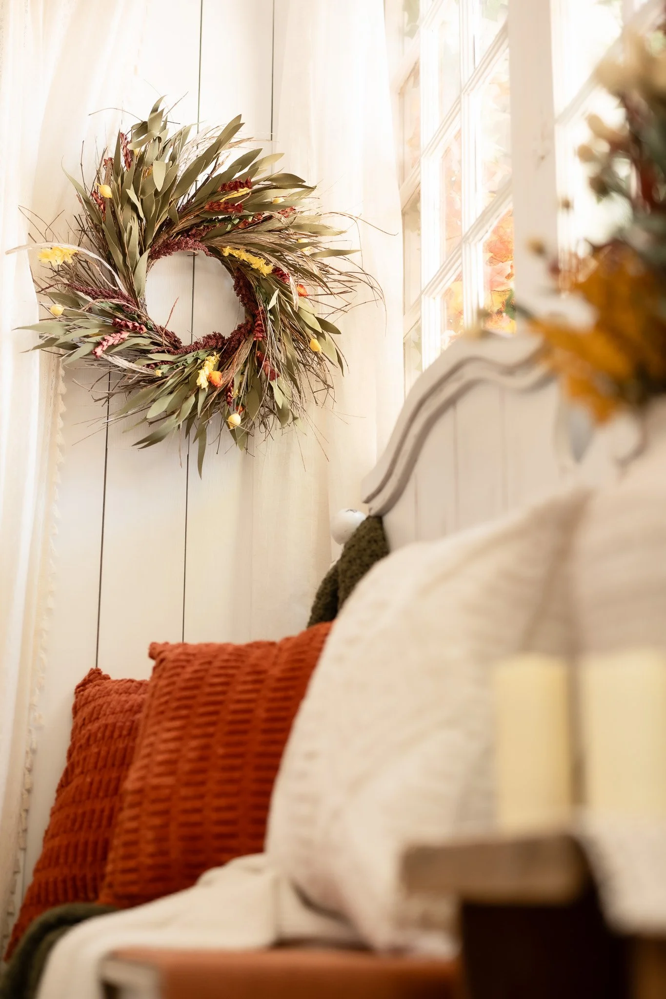 Close-up of a cozy, decorated indoor space with a white wooden bedframe and beige and burnt orange pillows, and a large autumn-themed wreath hanging on a wall near a window with natural light.