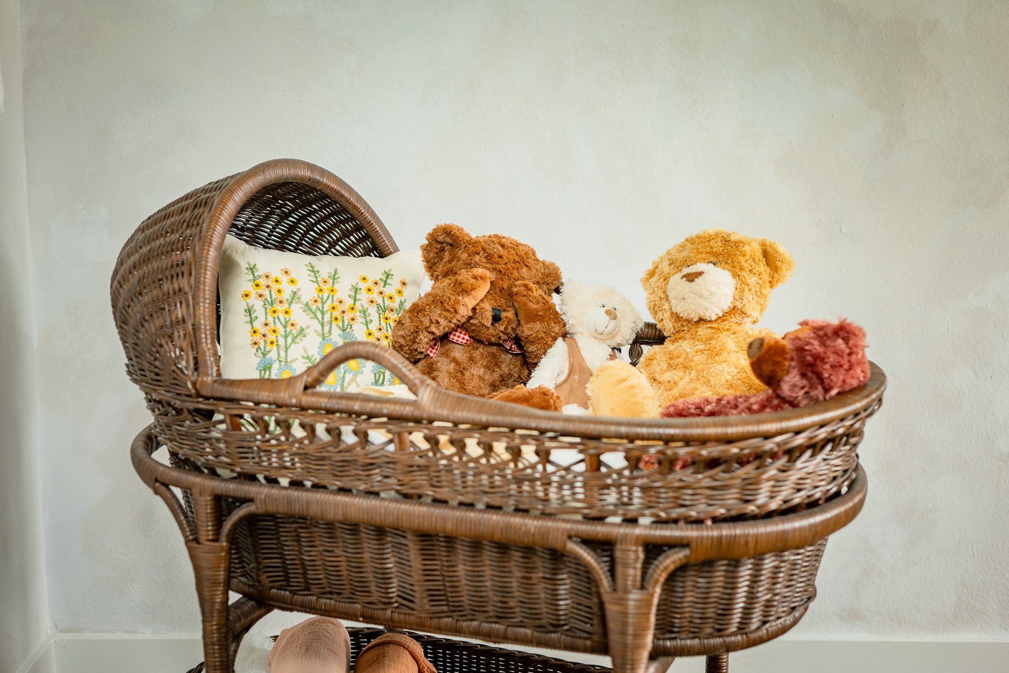 A wicker bassinet filled with various teddy bears and a decorative pillow with yellow and green embroidery, against a plain light-colored wall.