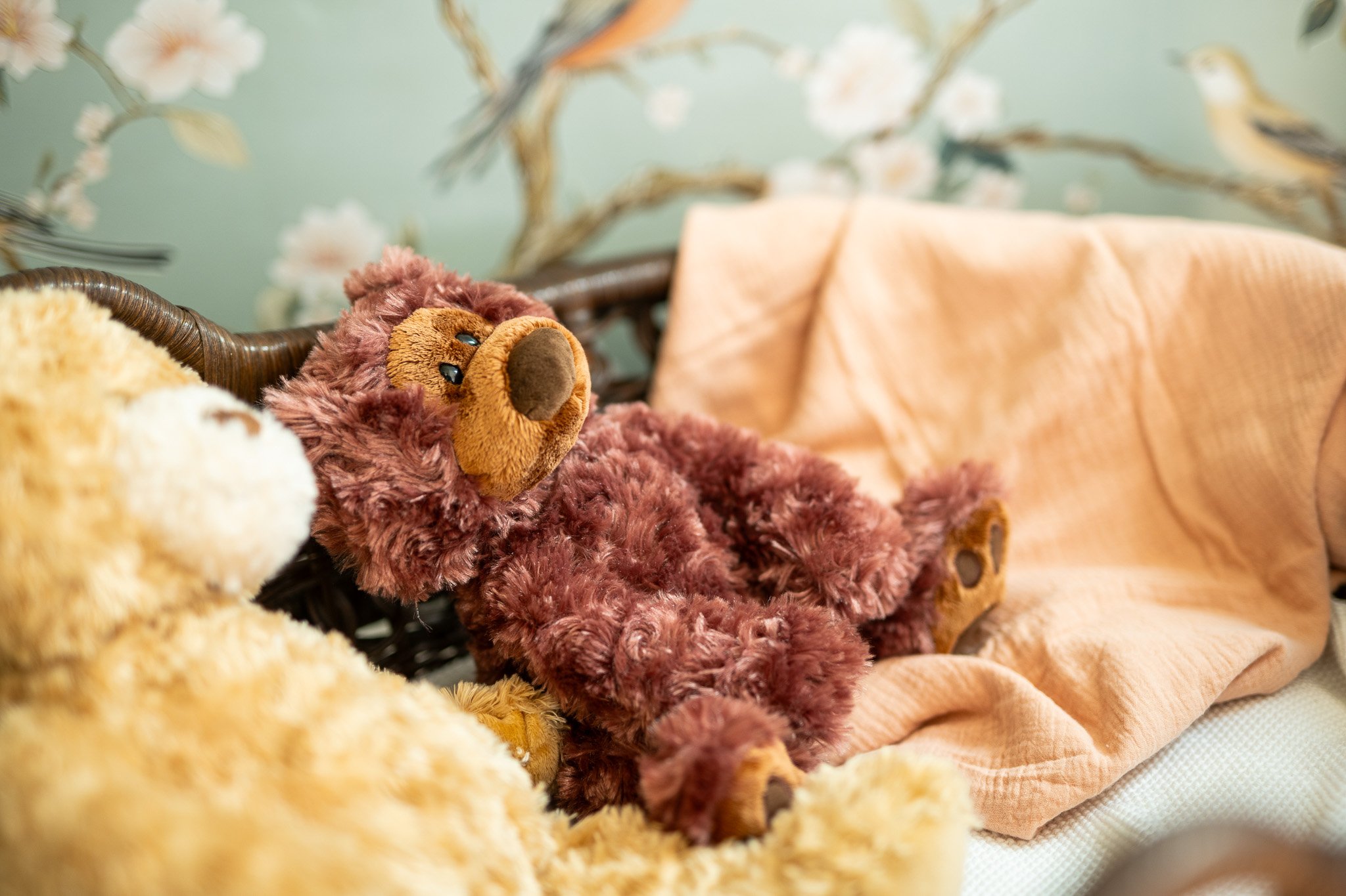 A plush teddy bear with reddish-brown fur and a darker brown nose, reclining on a woven brown basket with a peach-colored blanket draped behind it. The background features a floral wallpaper with birds in a soft, pastel color scheme.