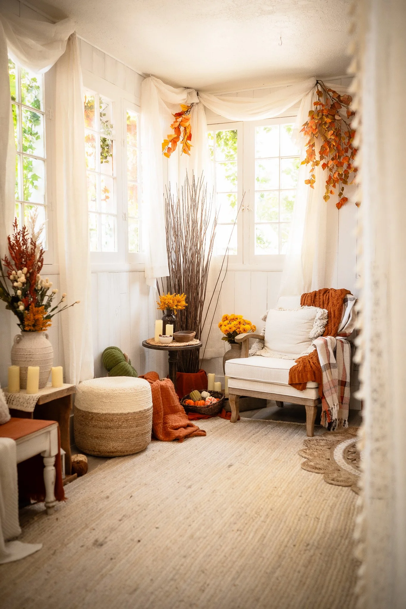 Cozy sunlit corner room with white walls, large windows, white curtains, autumn-colored leaves, a white armchair with a plaid blanket, decorative pillows, a small round table with candles and flowers, a pouf, and fall decorations including pumpkins a