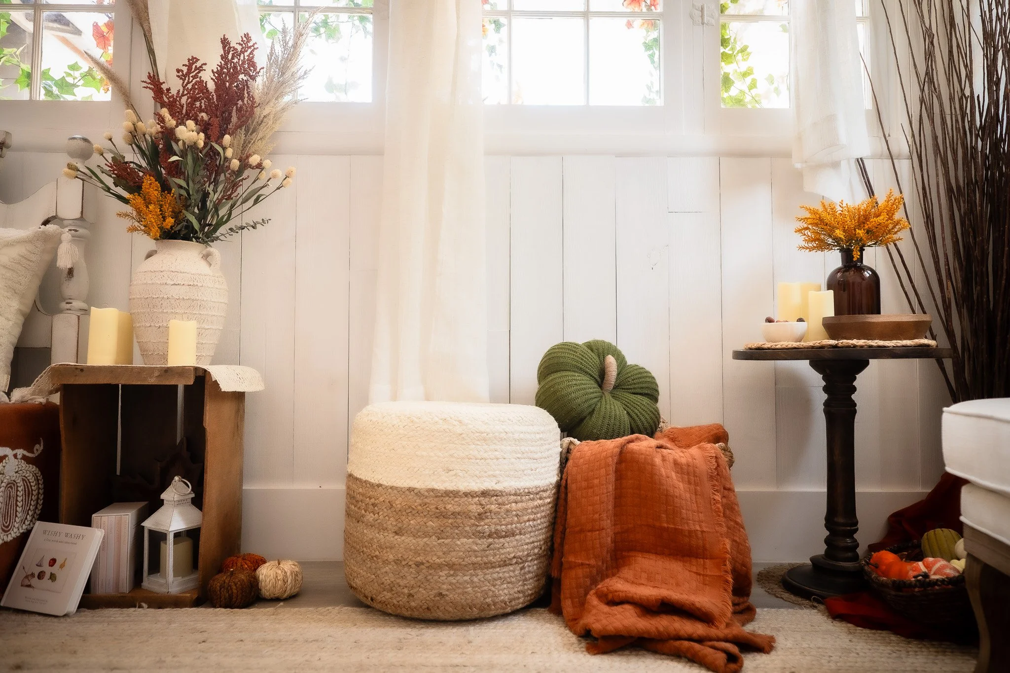 Cozy living room corner with white wooden walls, a large window with light curtains, and various decorative items including vases with dried flowers, candles, a woven ottoman, a green knotted pillow, orange blankets, a small crate with books and pump