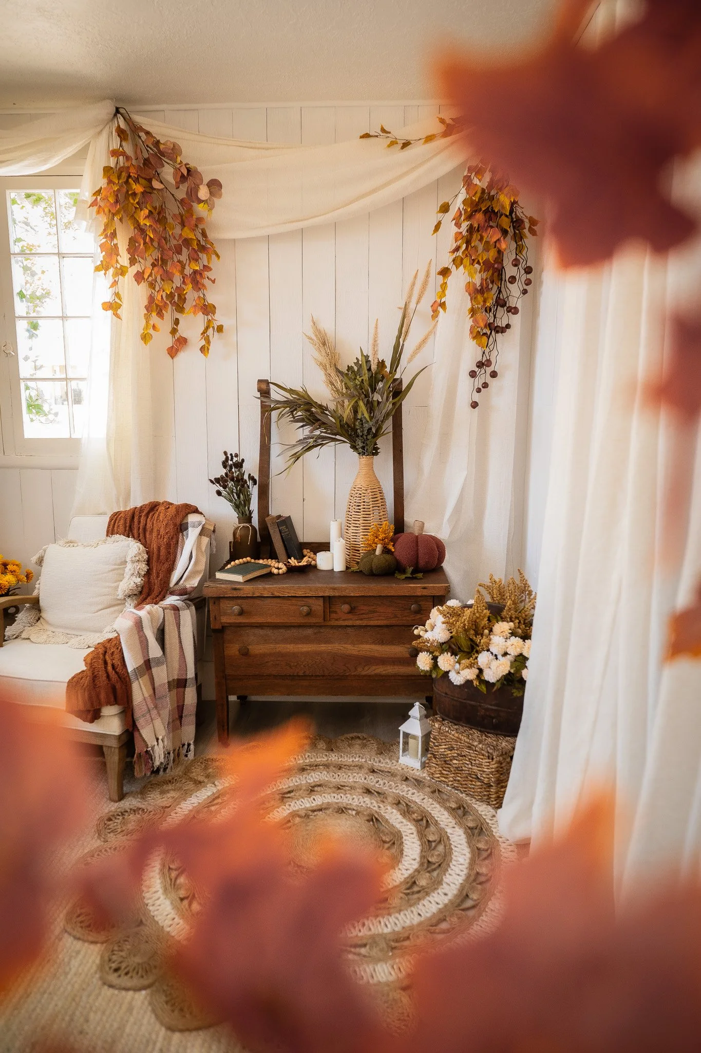 Cozy living room corner decorated for fall with orange and yellow leaves, a wooden cabinet with autumn-themed decor, a wreath with fall leaves on the wall, a white lantern on the floor, and a circular braided rug.