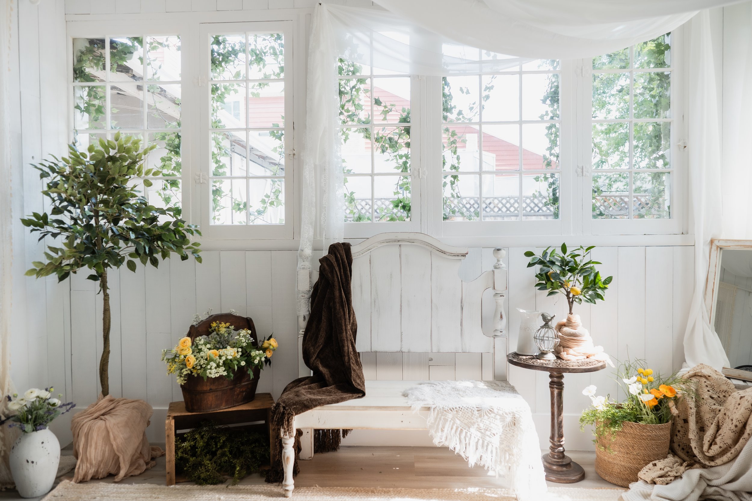 Bright room with white wood-paneled walls, multiple large windows with white curtains, and outdoor greenery visible outside. Small table with plants and flowers, white wooden bench with draped fabrics, and decorative items like vases and flowers.