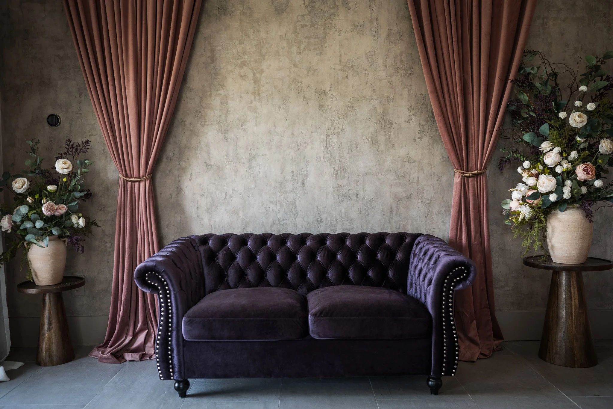 A purple velvet tufted sofa with nailhead trim, flanked by two wooden side tables with large floral arrangements, in front of a textured beige wall with pink curtains on either side.