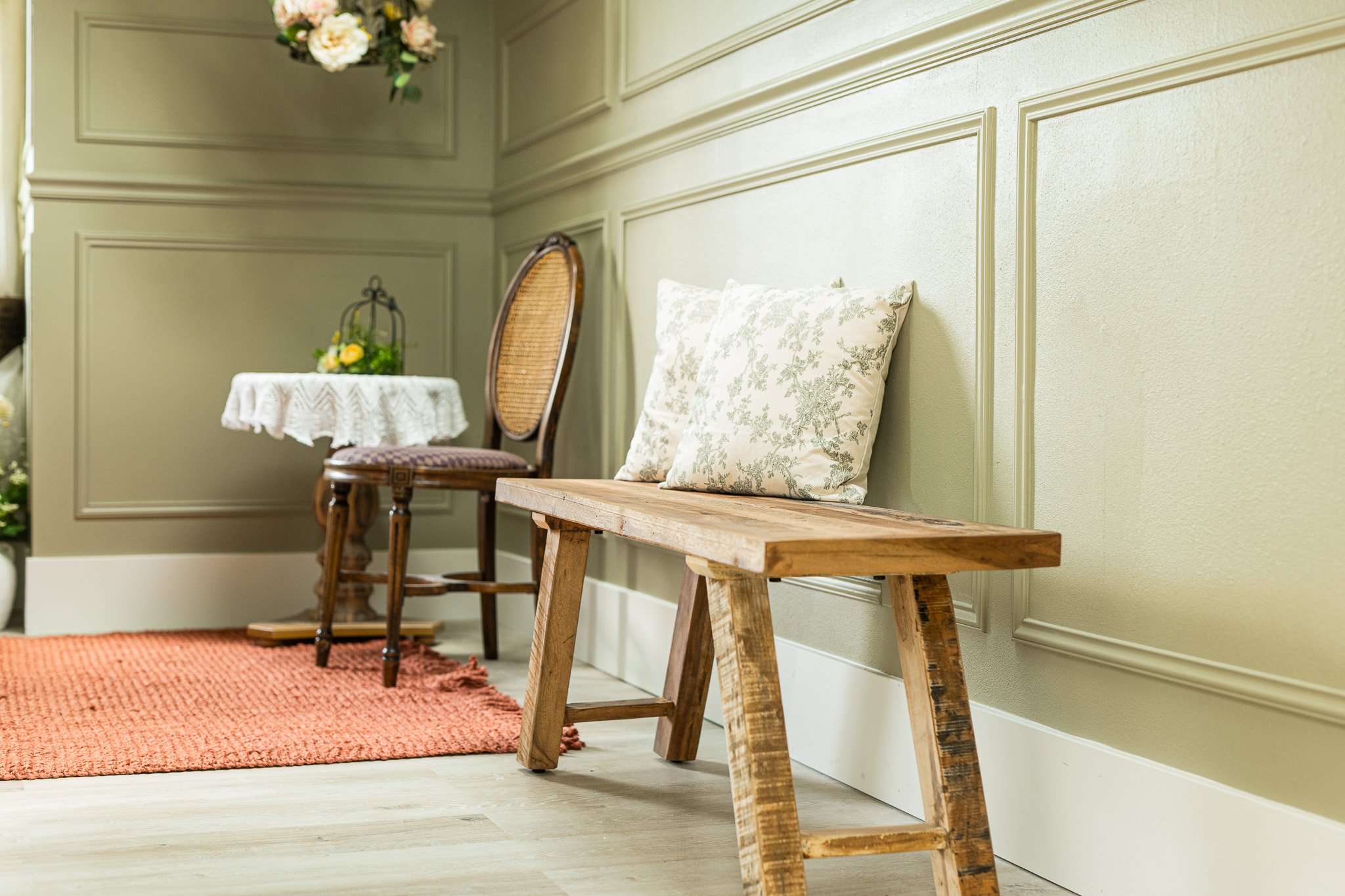 Interior of a room with a wooden bench with pillows, a vintage chair, and a small table with floral decoration, on a reddish-orange rug and light green walls.